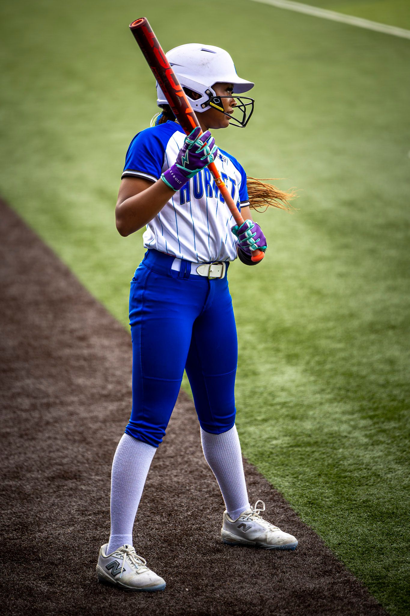 A softball player in a blue and white uniform and batting helmet holds a red bat while waiting on the field.