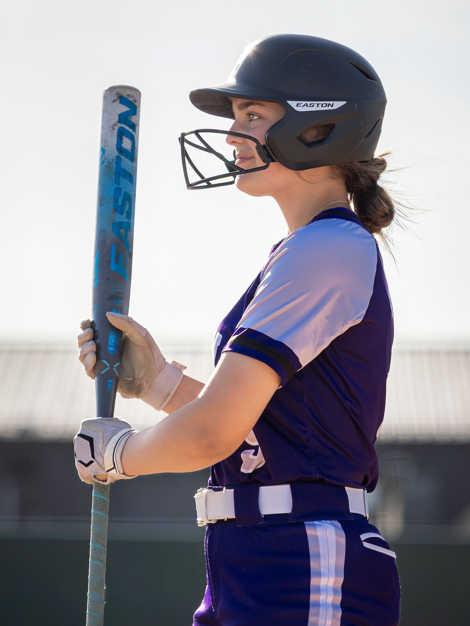 Softball player in a purple uniform and Easton batting helmet with face guard holds an Easton bat while waiting on deck.