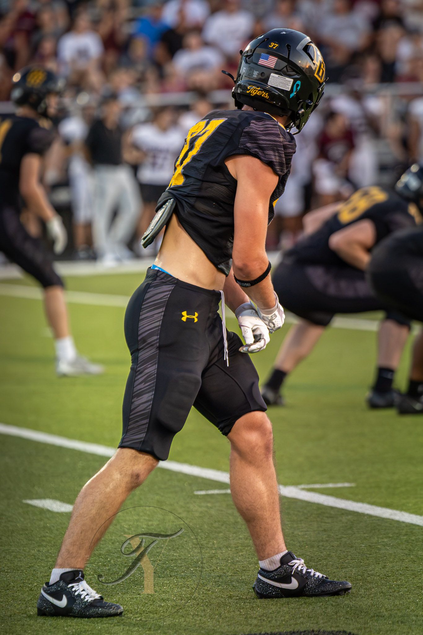 Football player in black and gold uniform with Under Armour gear stands on turf field during a game.