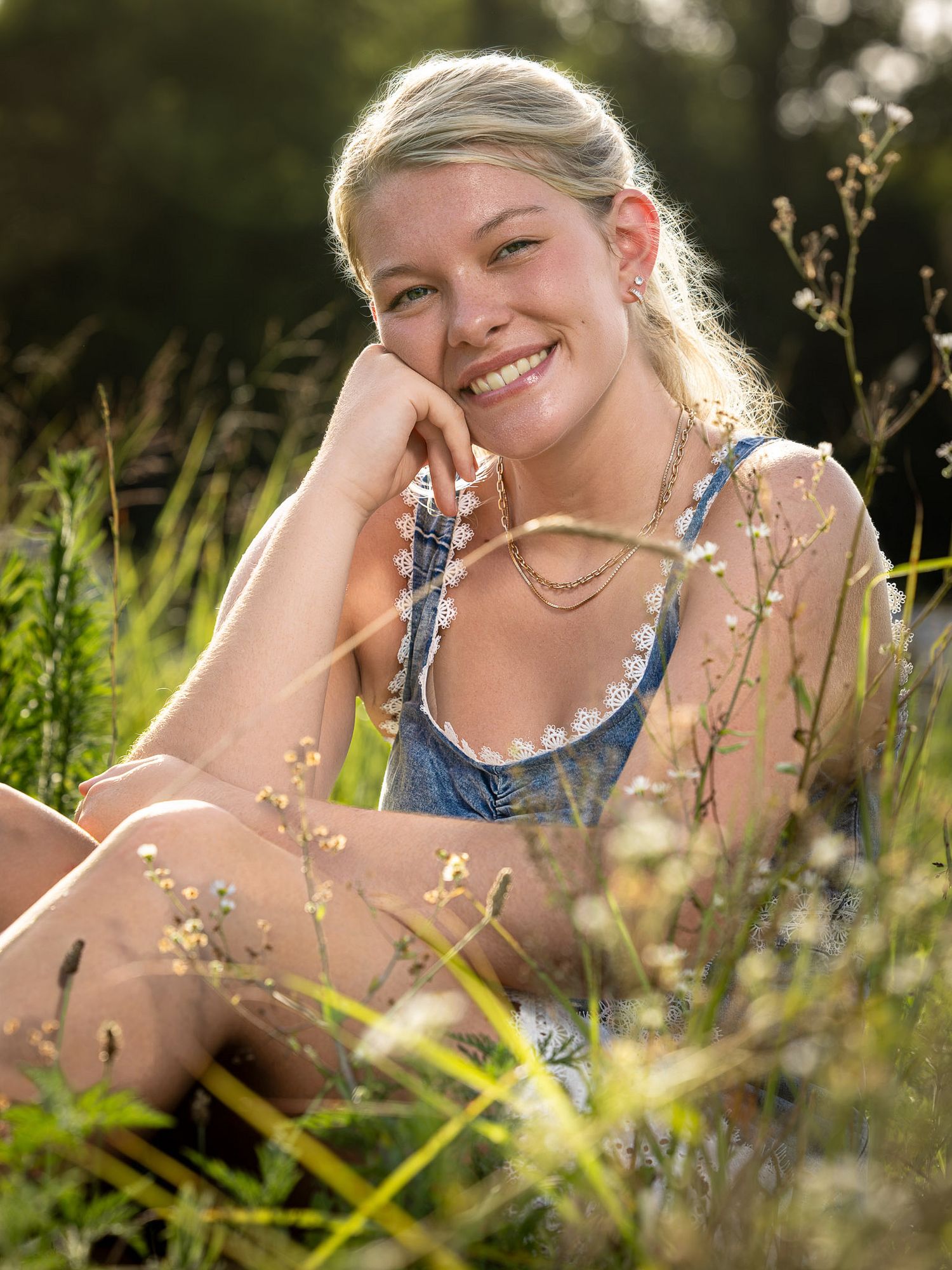 Elkins high school senior sitting in a sunny meadow wearing a denim top, surrounded by wildflowers and tall grass in West Fork