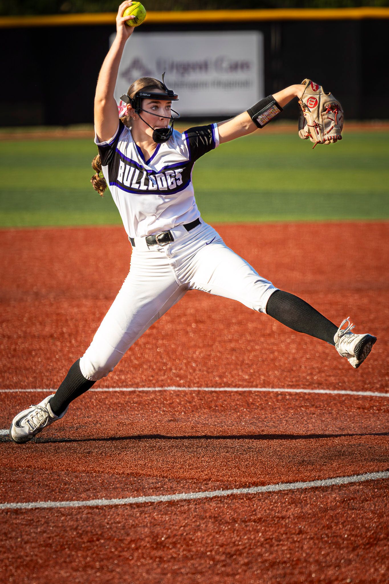 Softball pitcher in a white Bulldogs jersey winds up mid-delivery, holding the ball high with her glove extended on the red dirt.