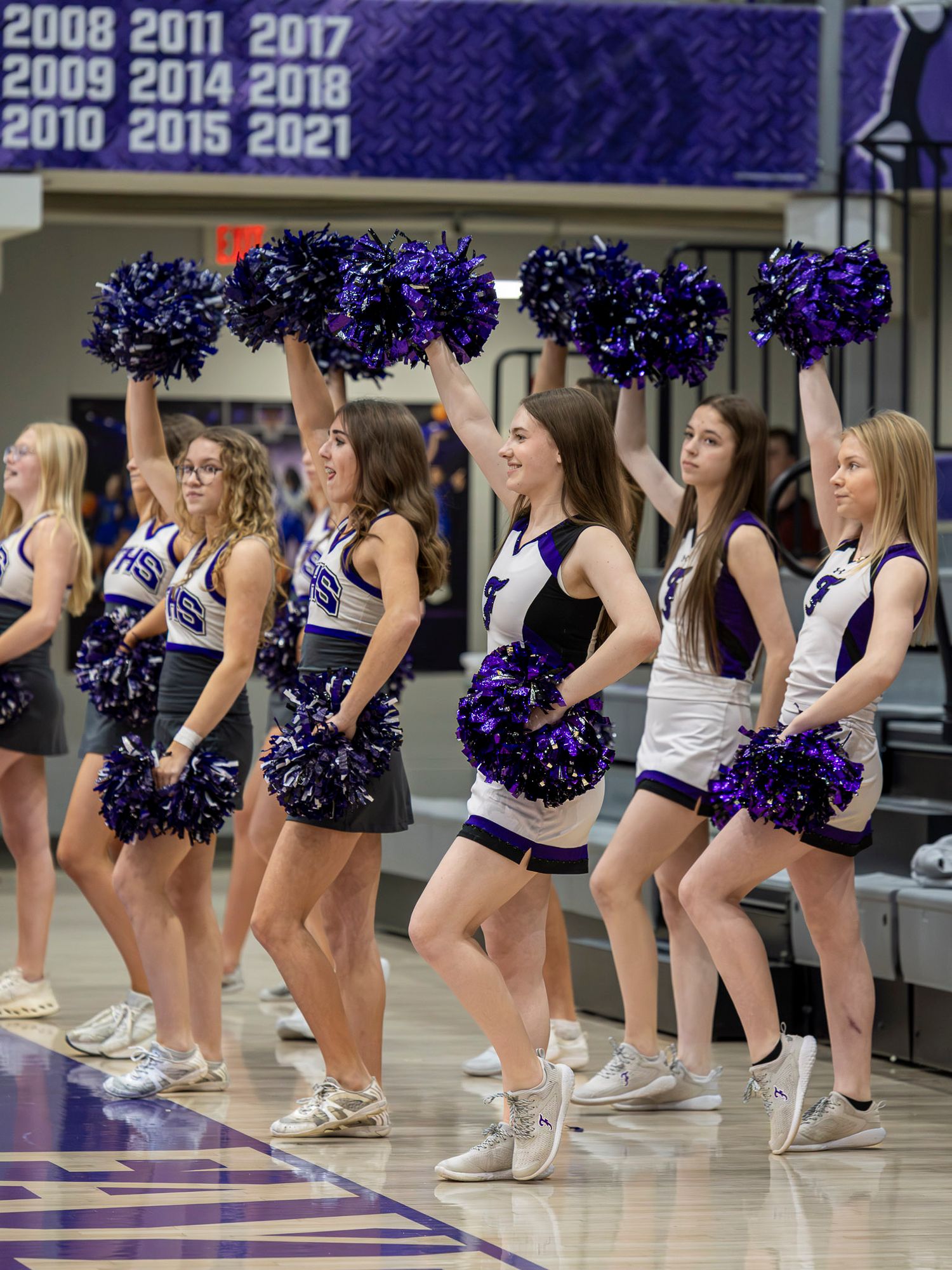 College cheerleaders in purple and white uniforms perform synchronized dance routine during basketball game.