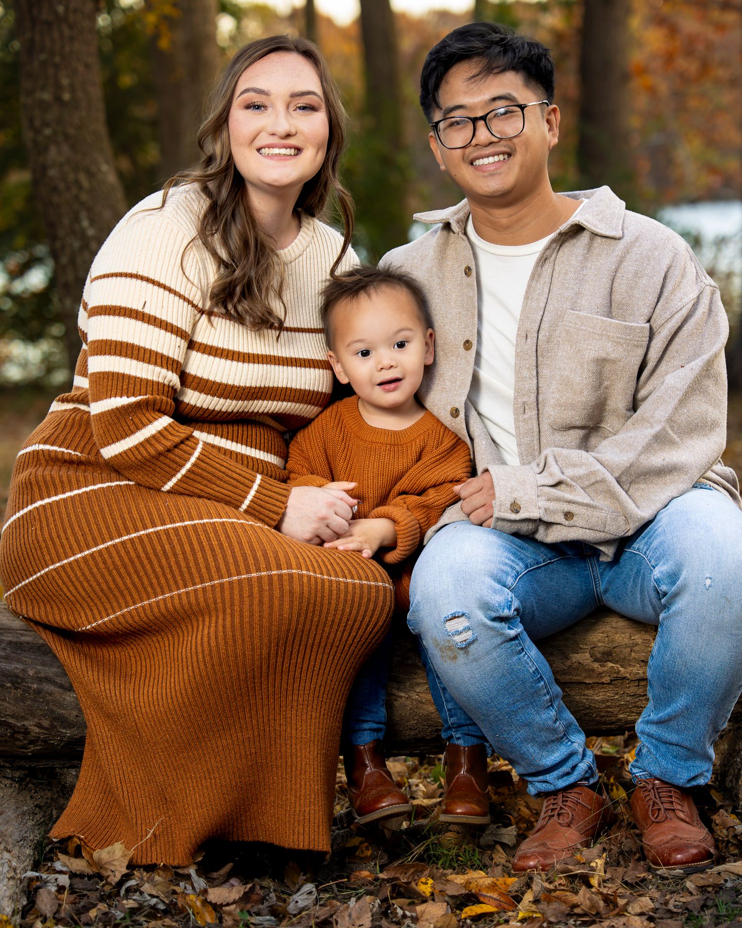 A young family of three poses outdoors in autumn, wearing warm earth-toned outfits, with a smiling toddler between his parents.