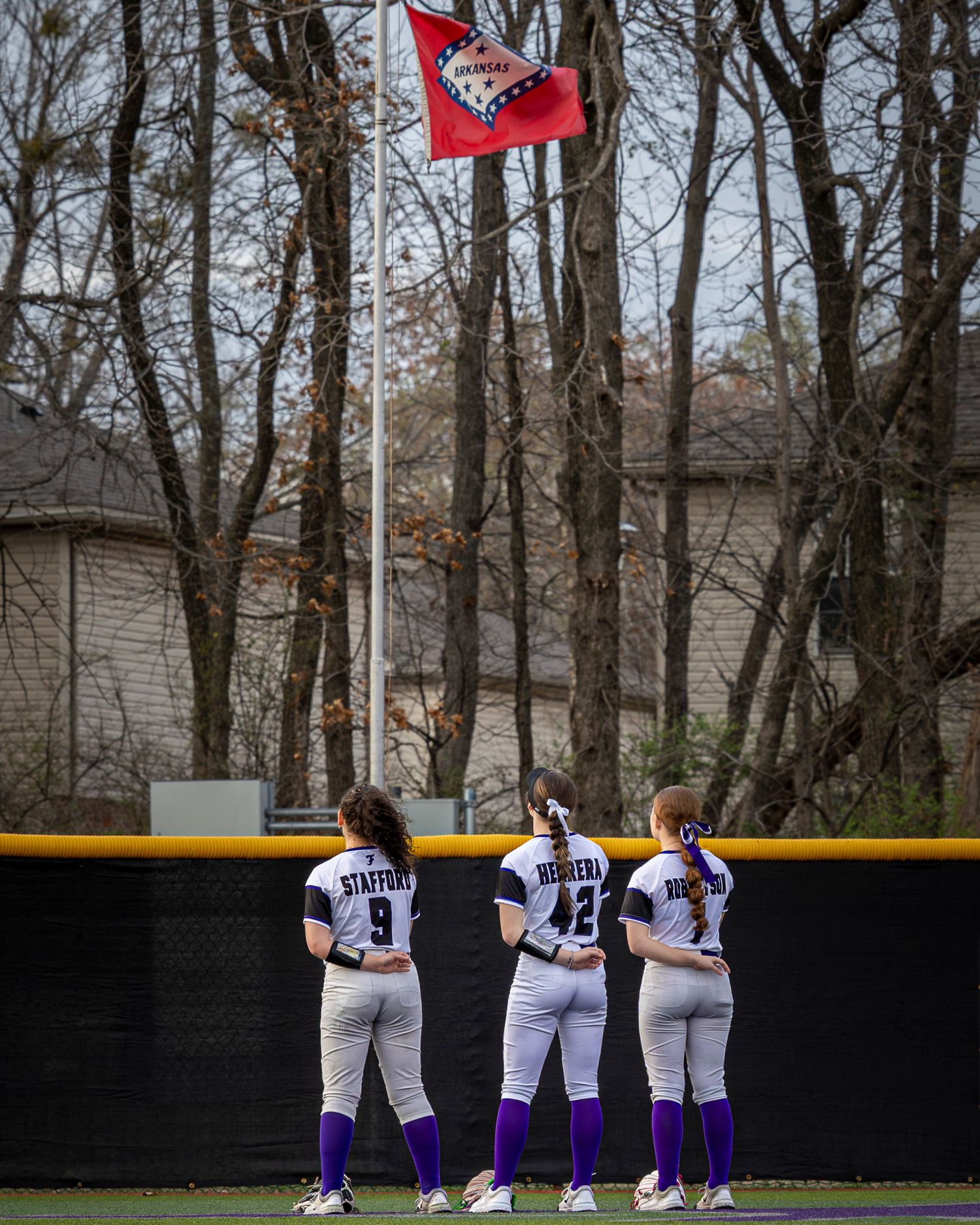 Three softball players in white jerseys and purple socks stand facing a flag on a pole during the national anthem before a game.