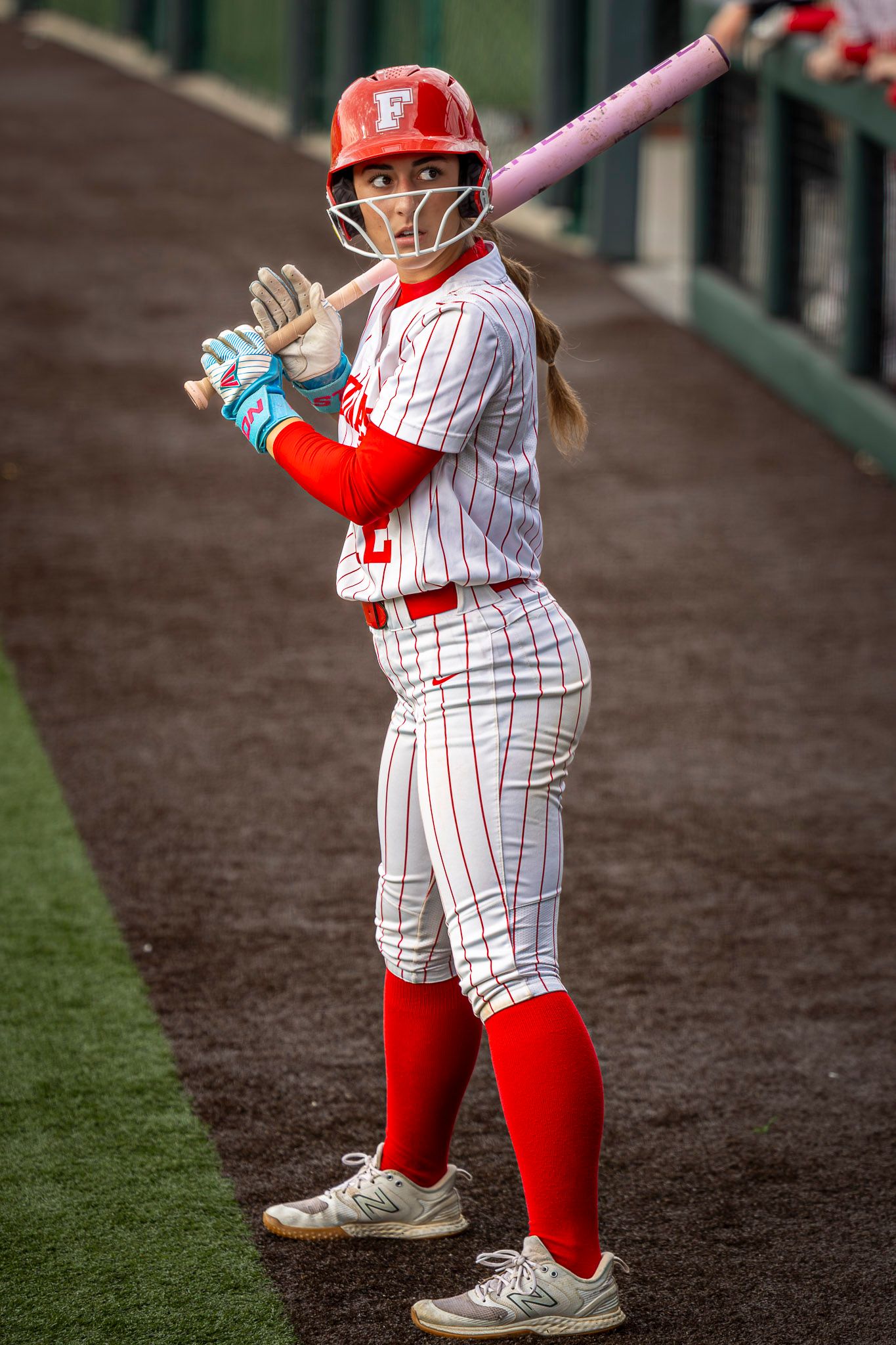 Softball player in a red helmet, pinstriped white uniform and red socks holds a pink bat on her shoulder while waiting in the on-deck area.