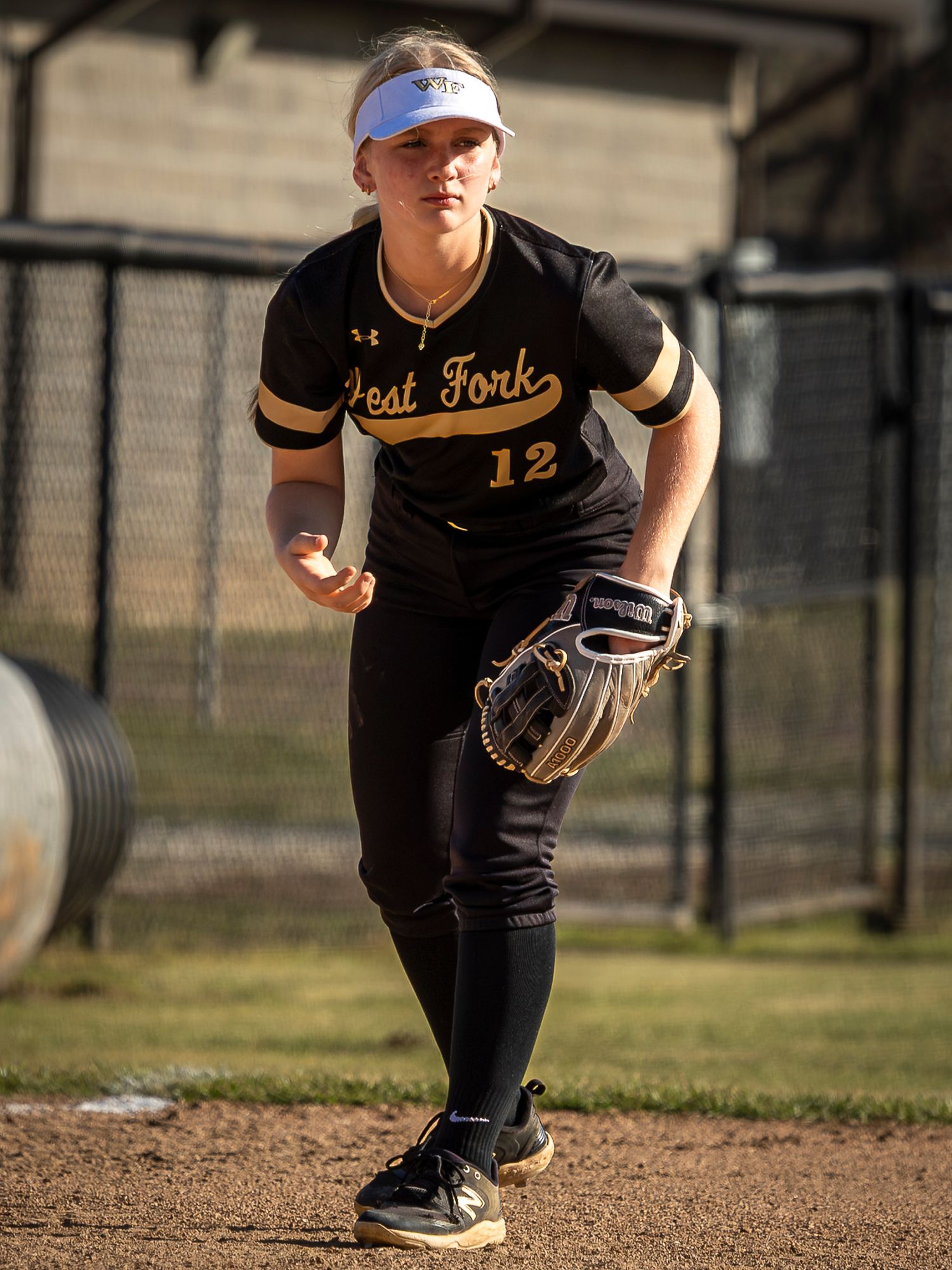 West Fork softball player in black number 12 jersey, visor and glove crouches ready in the infield during a game.