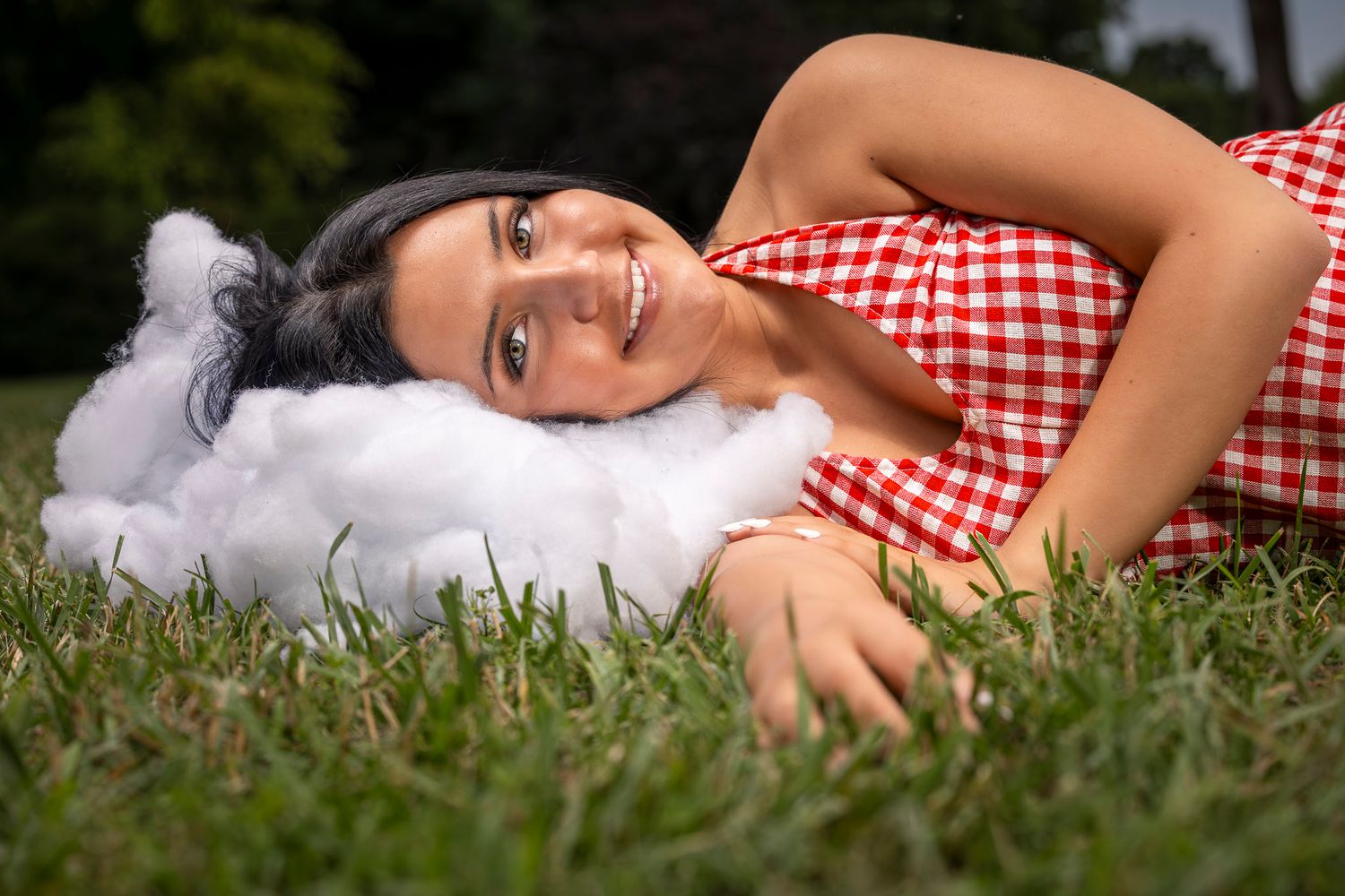 Elkins senior in red gingham dress lying on grass, smiling while resting head on a fluffy white cloud pillow outdoors.