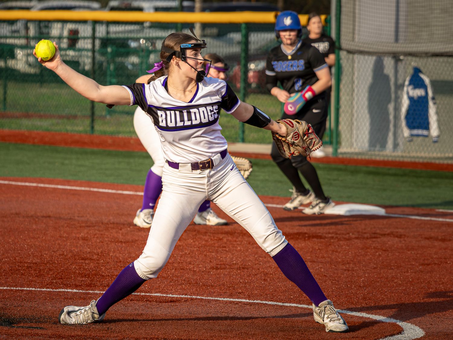 Bulldogs softball player in a white and purple uniform winds up to throw a yellow softball during a game on a dirt infield.