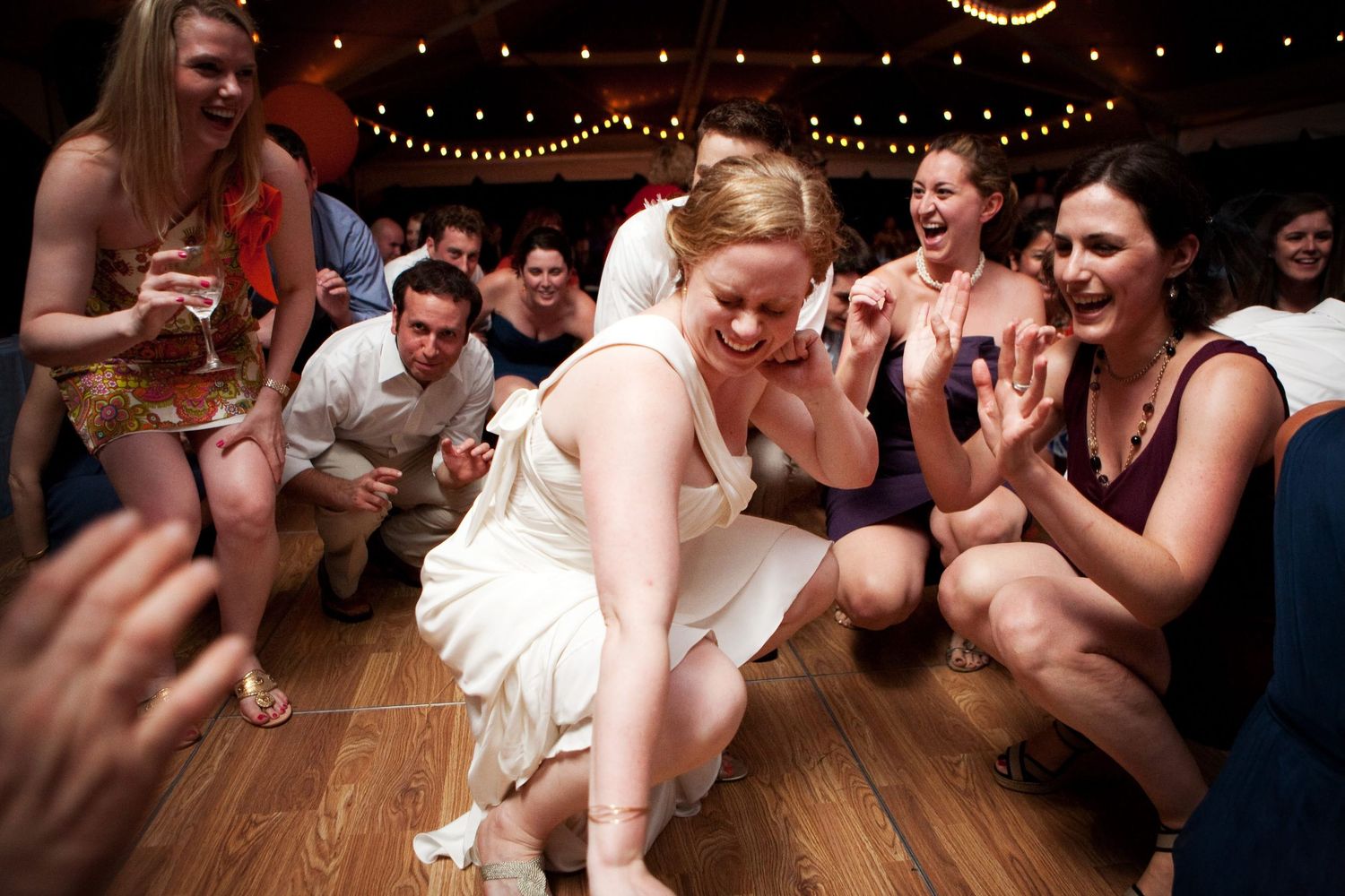 Party guests gather around someone in a white dress dancing on a wooden floor during a celebration with string lights overhead.