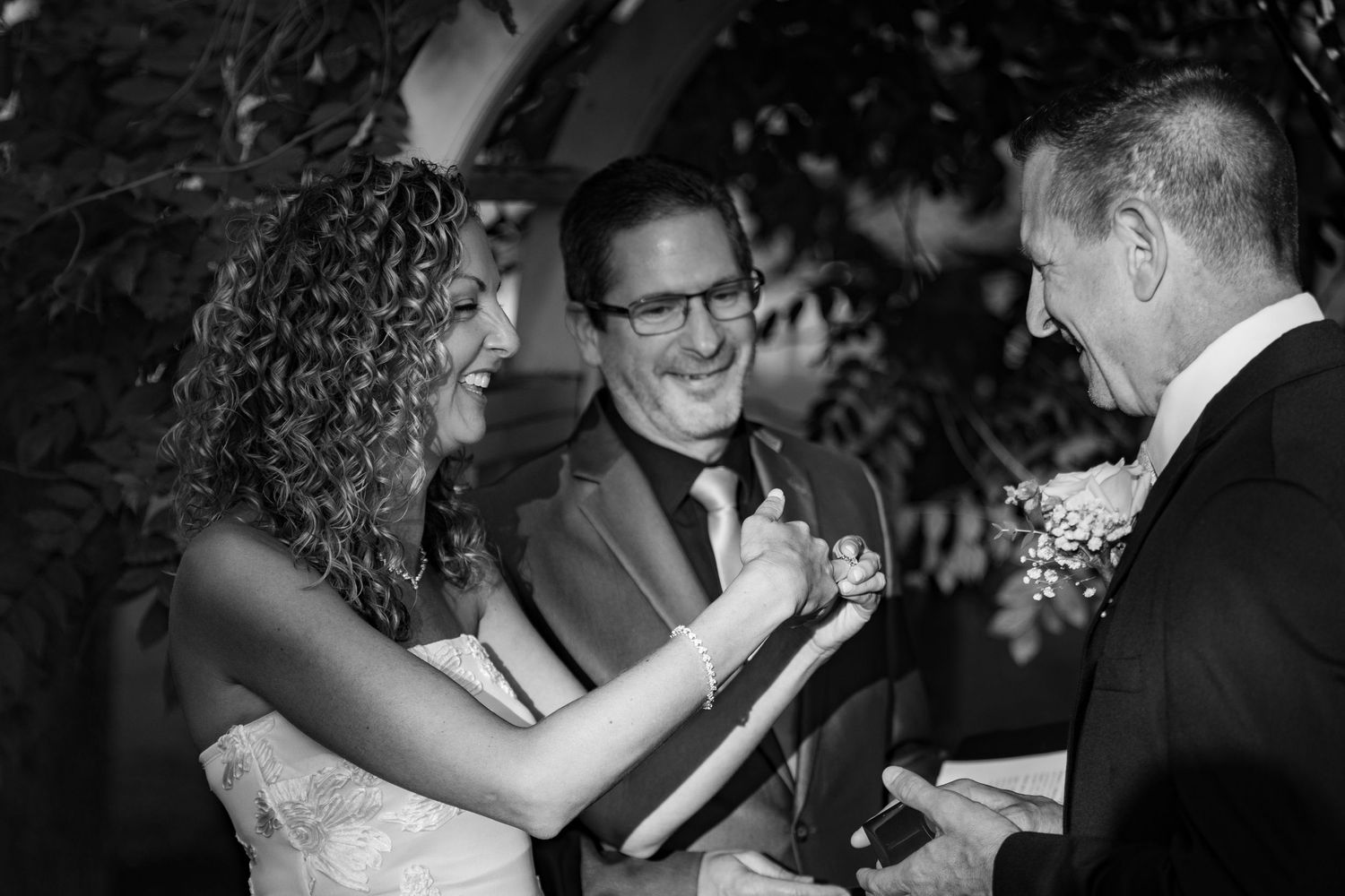 Black and white photo of a joyful bride and groom laughing with officiant during outdoor wedding ceremony.