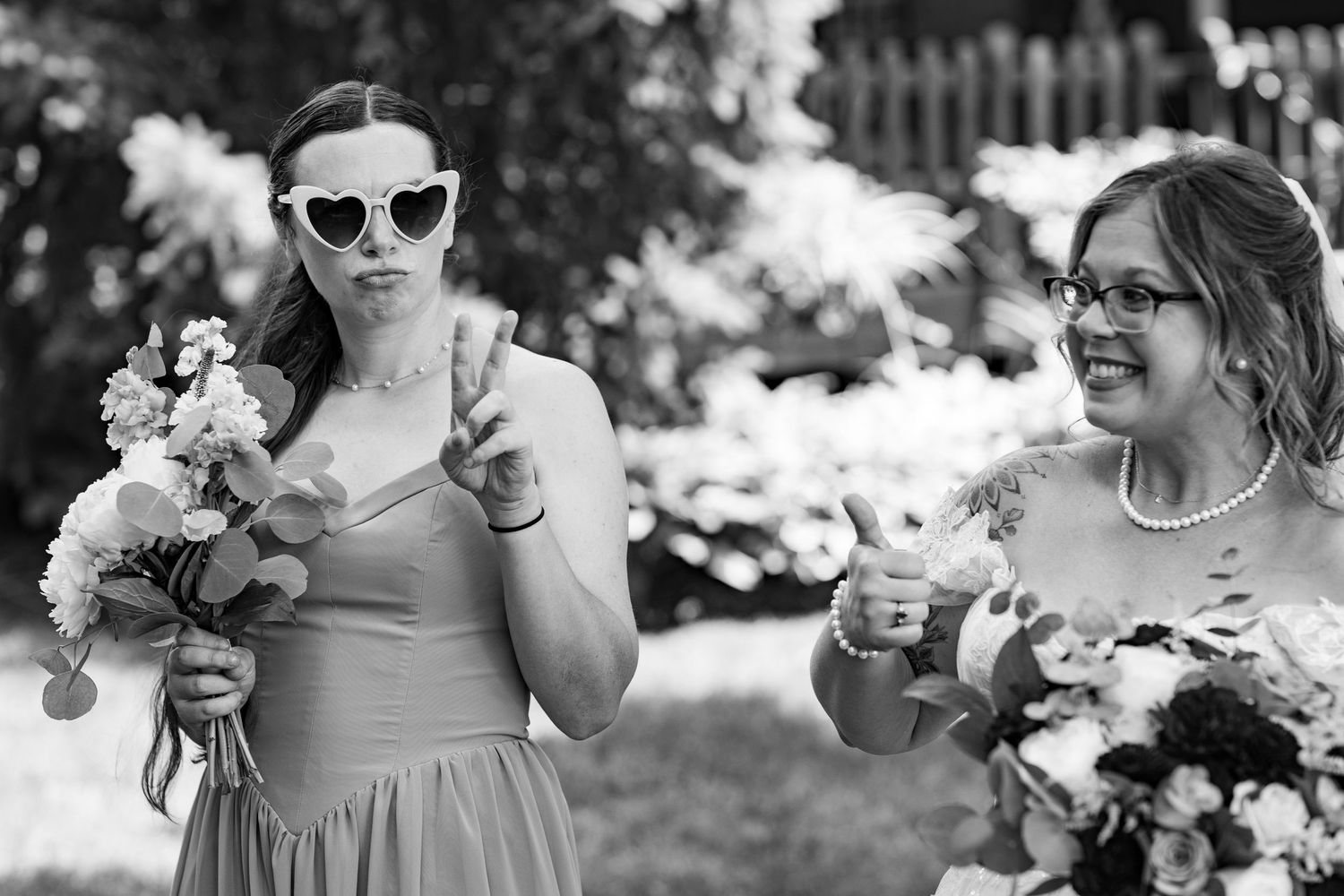 Two bridesmaids laughing outdoors, one wearing heart sunglasses and flashing a peace sign.