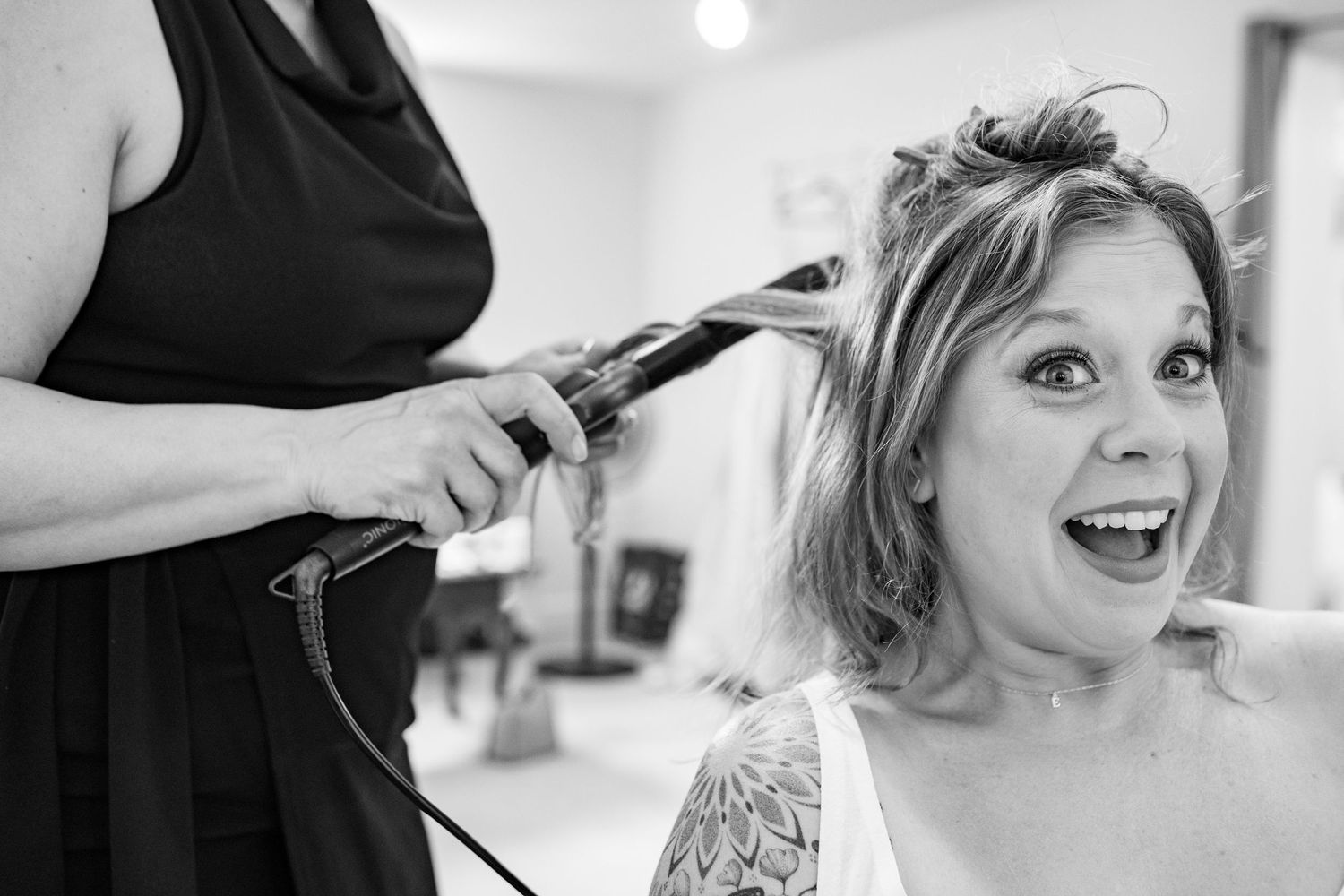 Bride laughing with joy as hairstylist curls her hair with a flat iron during wedding preparations.