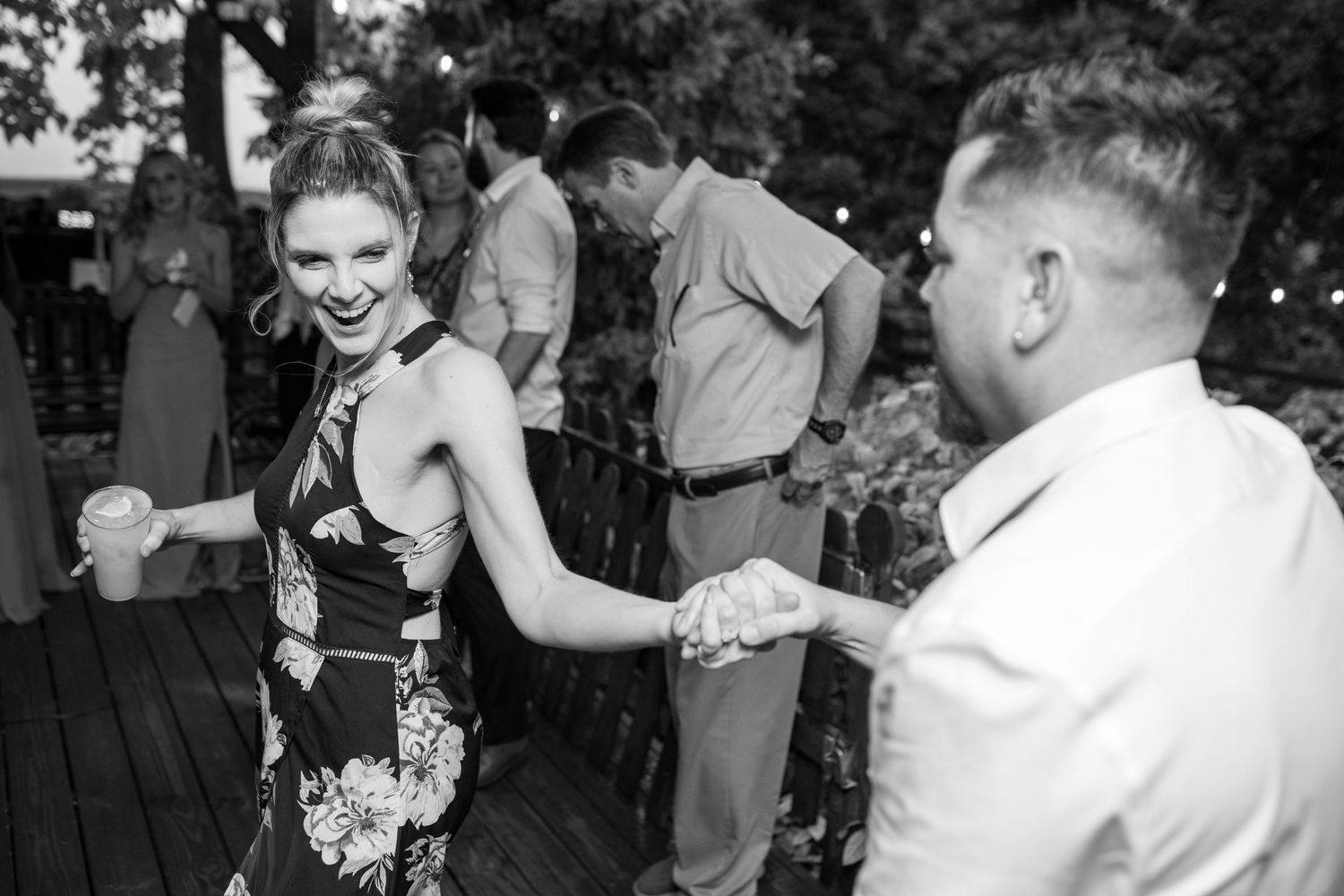 Black and white photo of a woman in floral dress laughing while dancing with a partner at a wedding.
