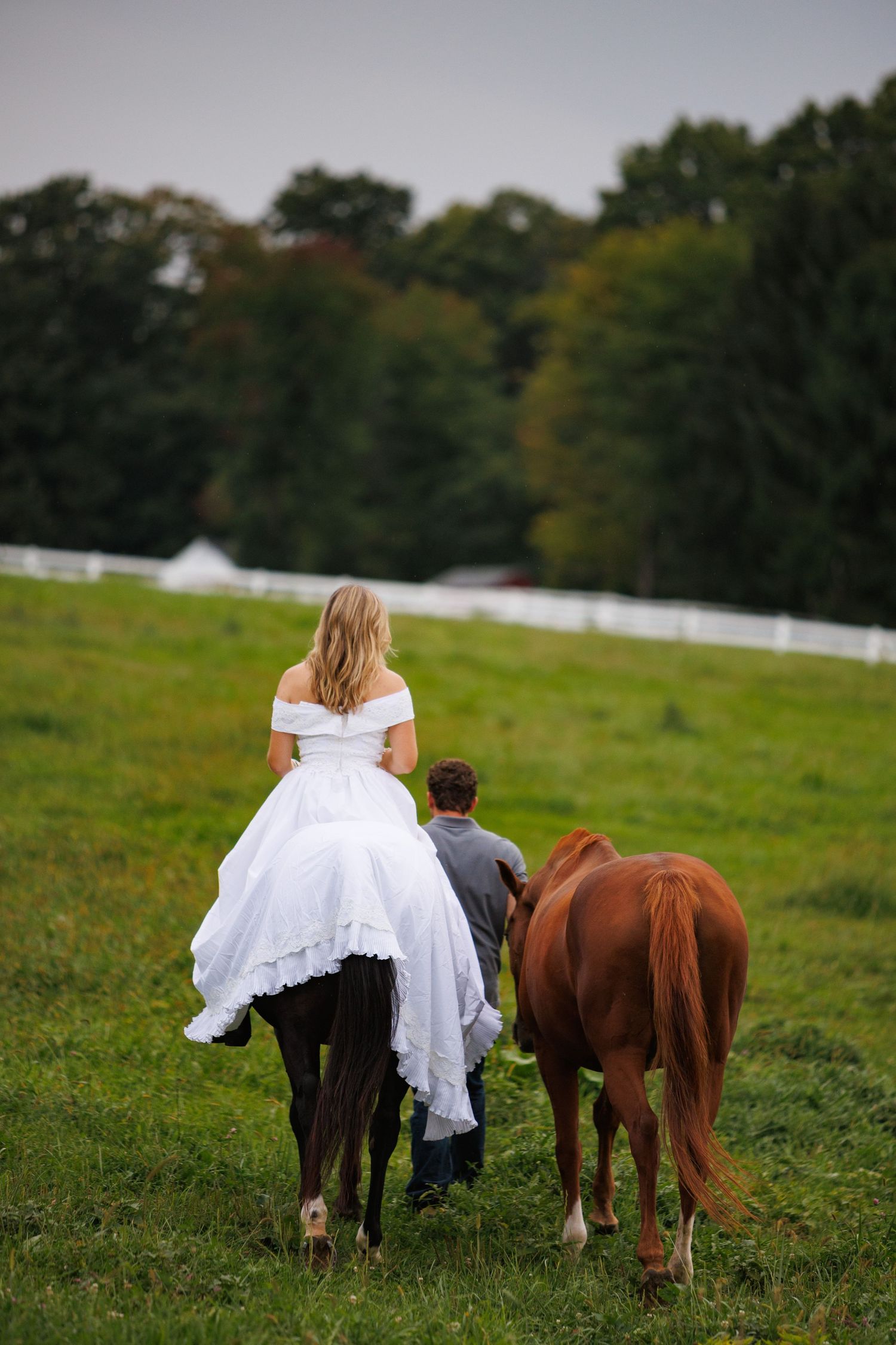 Bride in white gown rides dark horse as groom walks beside her with brown horse in green field.