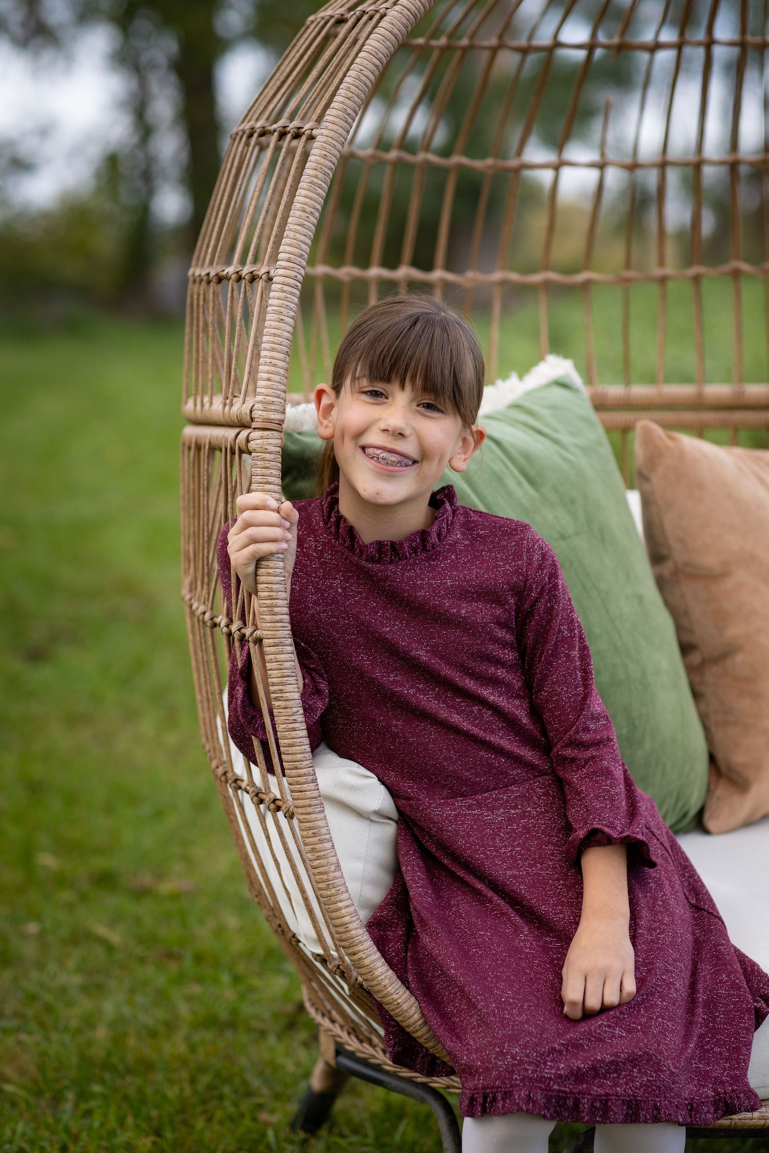 Child sitting in a rattan hanging chair with cushions on a grassy lawn wearing a burgundy dress.