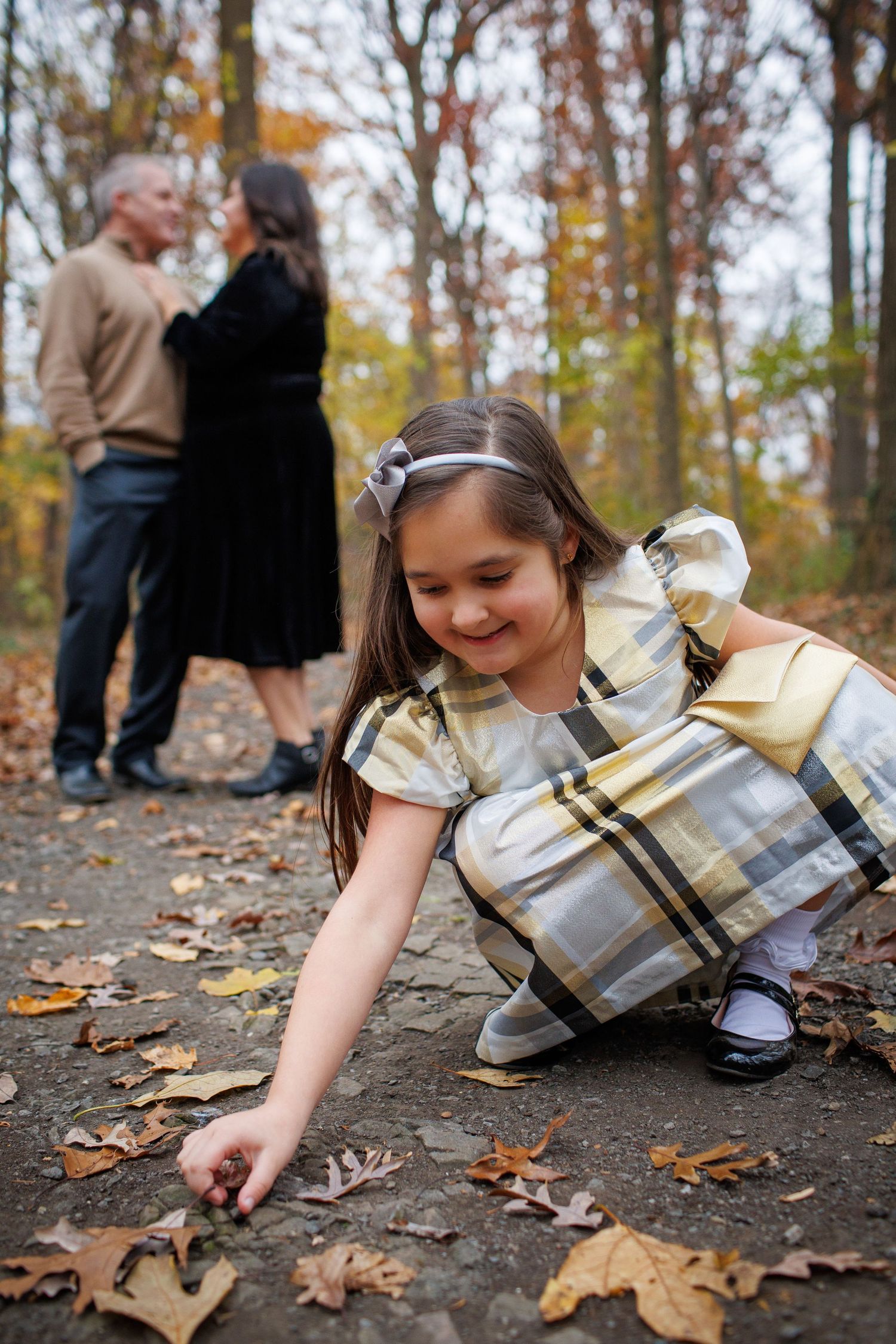 A young girl in a plaid dress crouches down to examine fallen autumn leaves while adults stand in background.