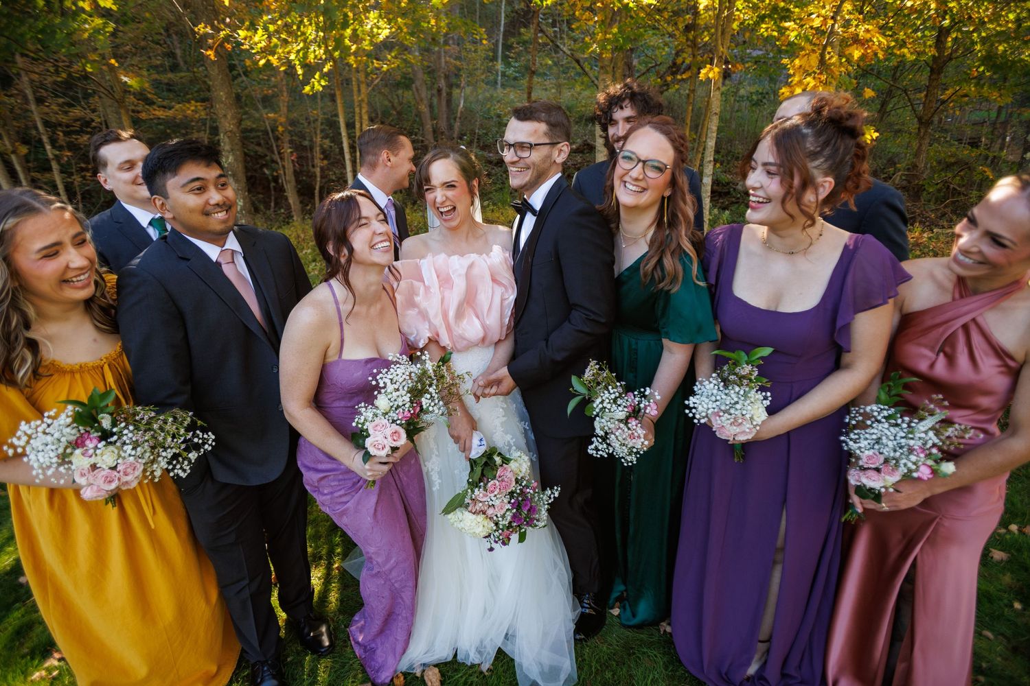 Wedding party smiling outdoors in autumn forest setting with bridesmaids in colorful dresses holding bouquets.