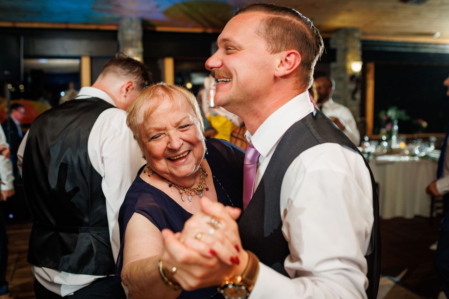 A groom in a vest laughs and dances with an older woman at a wedding reception.