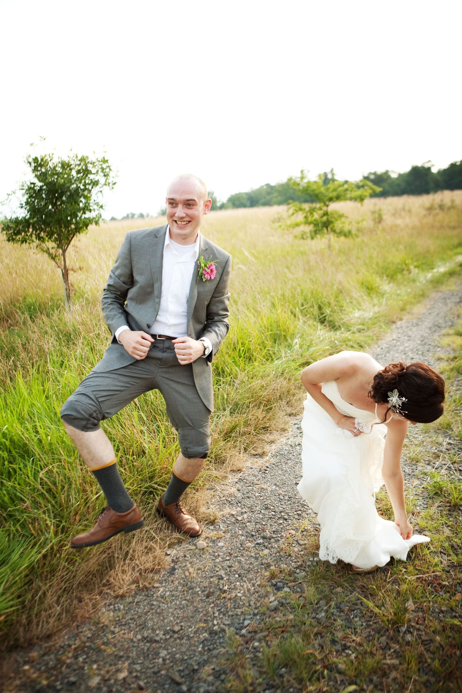 Playful newlyweds laugh together on a countryside path with tall grass.