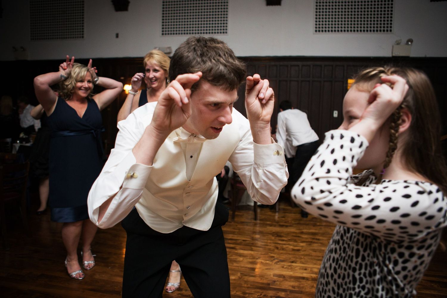 Party guests dancing and making exaggerated dance moves on a wooden dance floor at a celebration event.