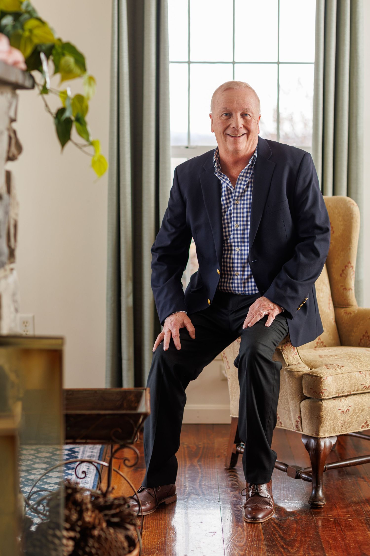 A person wearing a navy blazer and checkered shirt sits in an elegant room with hardwood floors and a beige armchair.