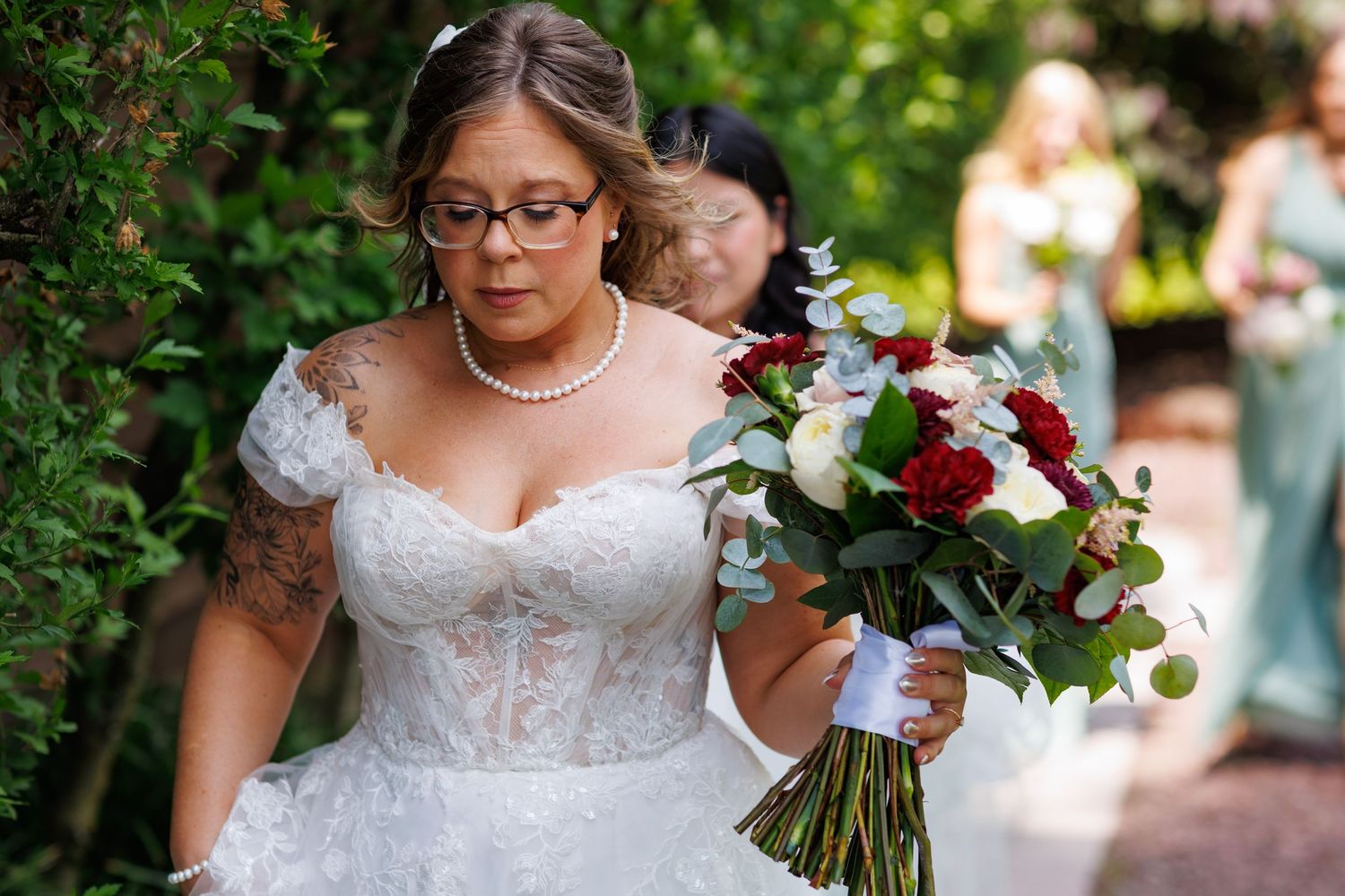 Bride in lace off-shoulder gown with glasses holding red and white bouquet walking down garden path.