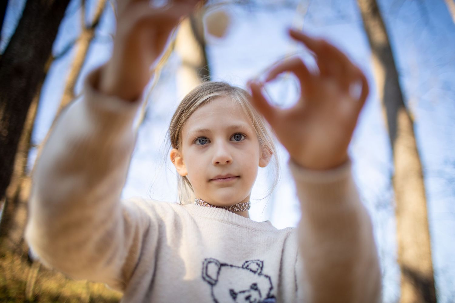 Child in a light sweater makes hand gestures against a blue sky in the woods.