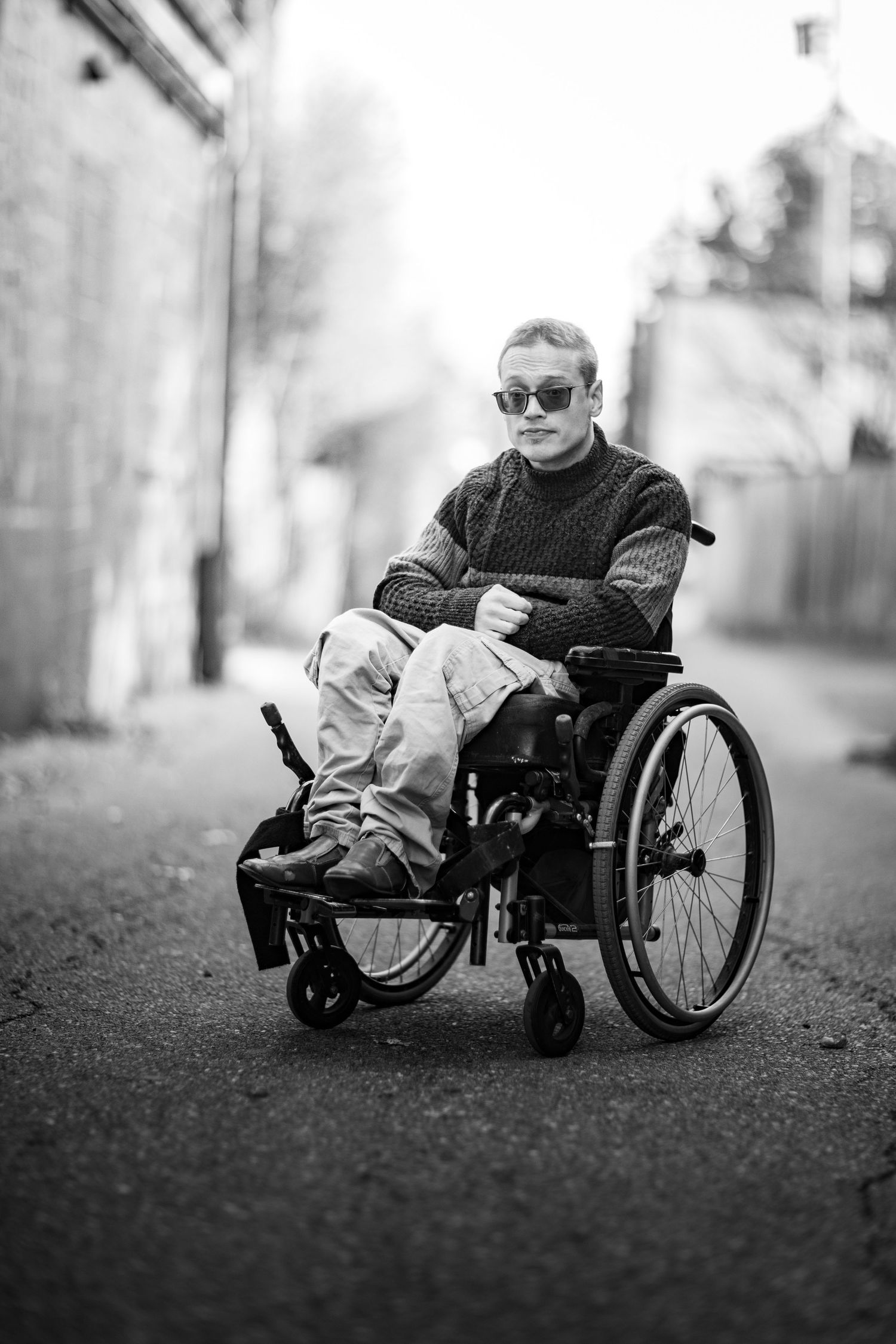 Black and white portrait of a person in a sweater and sunglasses seated in a wheelchair outdoors.