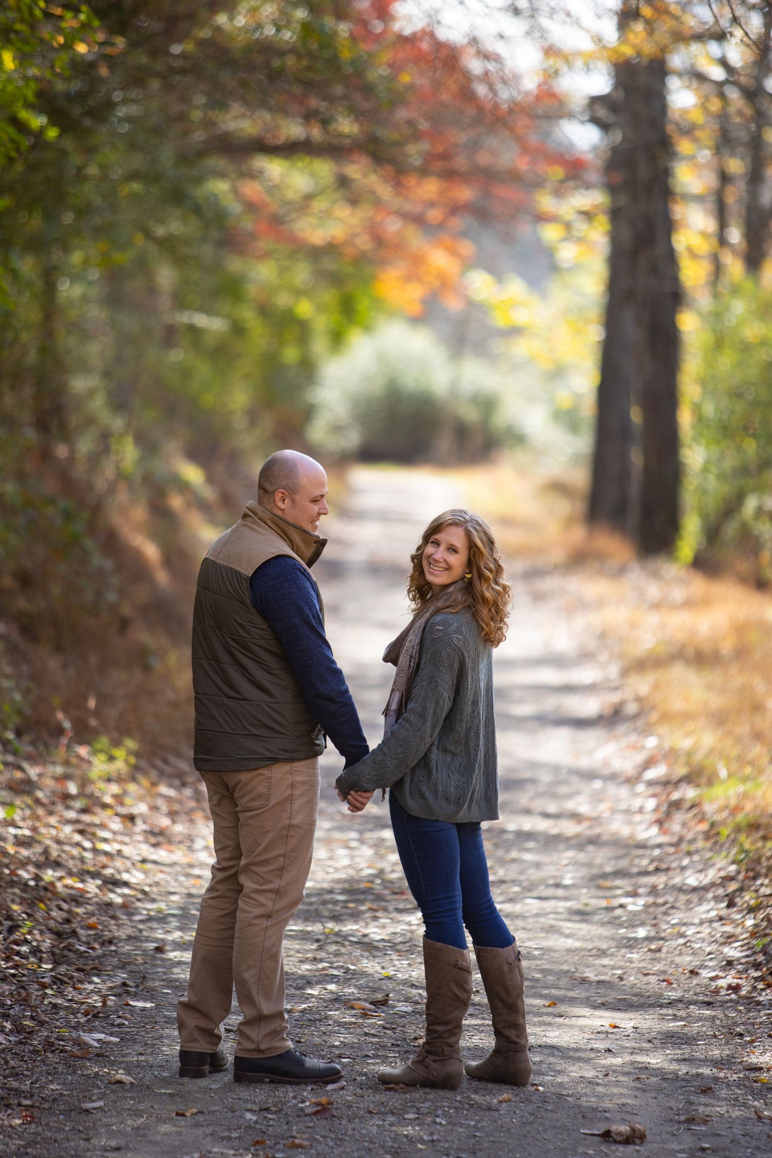 Two people share a tender moment on a leaf-covered autumn path surrounded by colorful fall foliage.