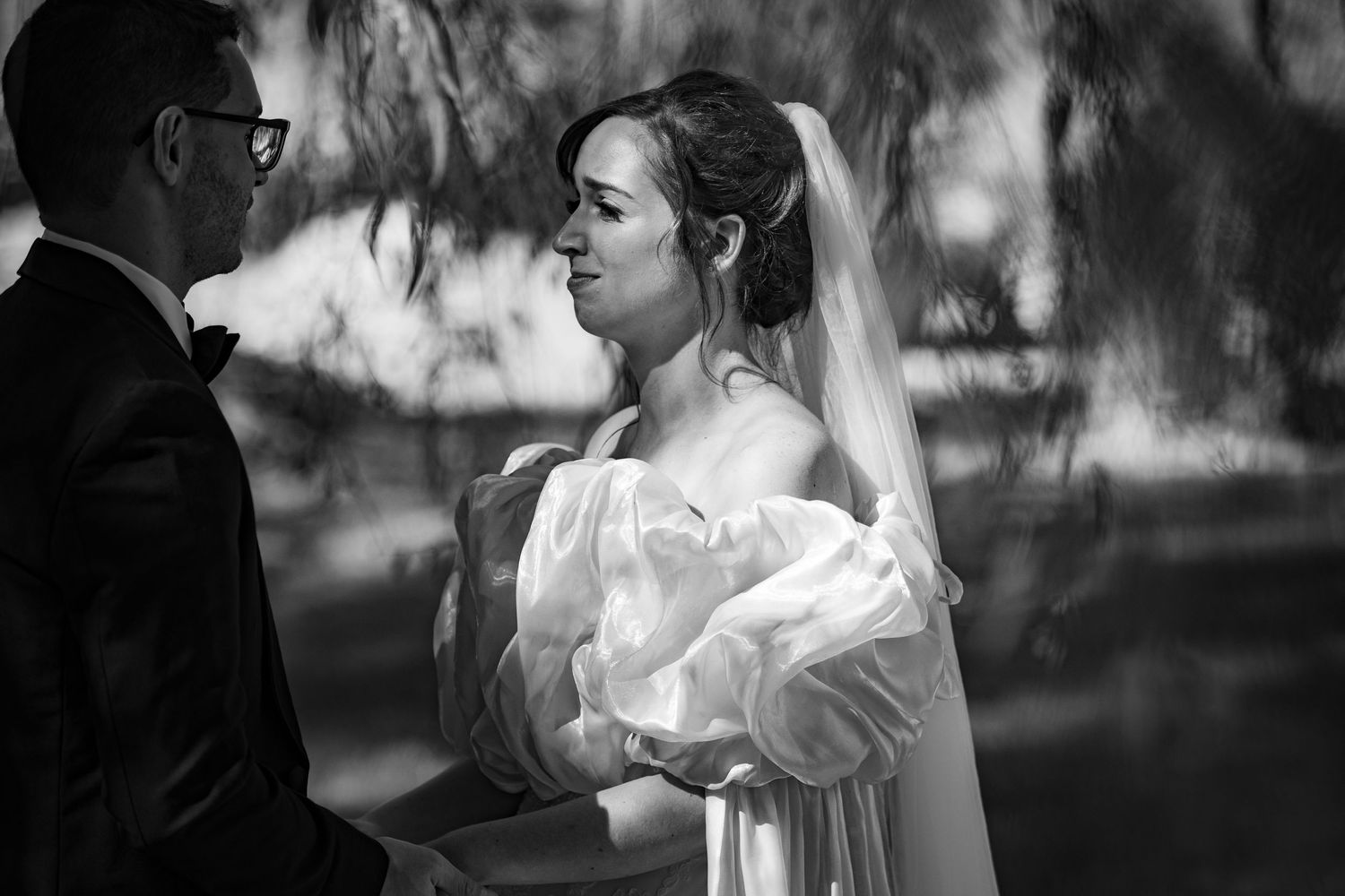 Elegant bride in off-shoulder gown and veil gazes thoughtfully outdoors in black and white portrait.
