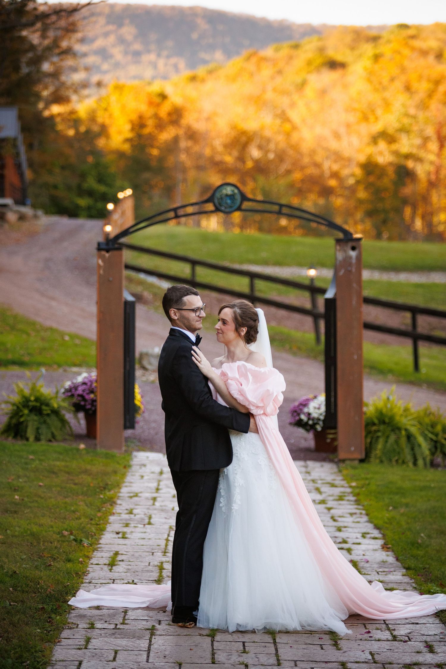 Newlyweds embrace under rustic wooden arch at sunset with colorful autumn mountain backdrop.