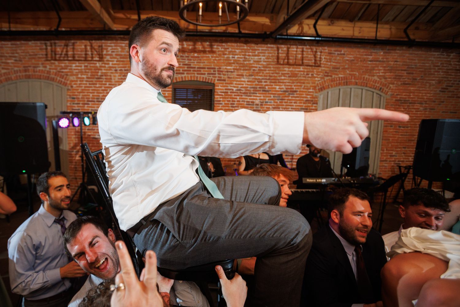 Groom celebrates at wedding reception, pointing excitedly while being lifted by guests in a brick-walled venue.