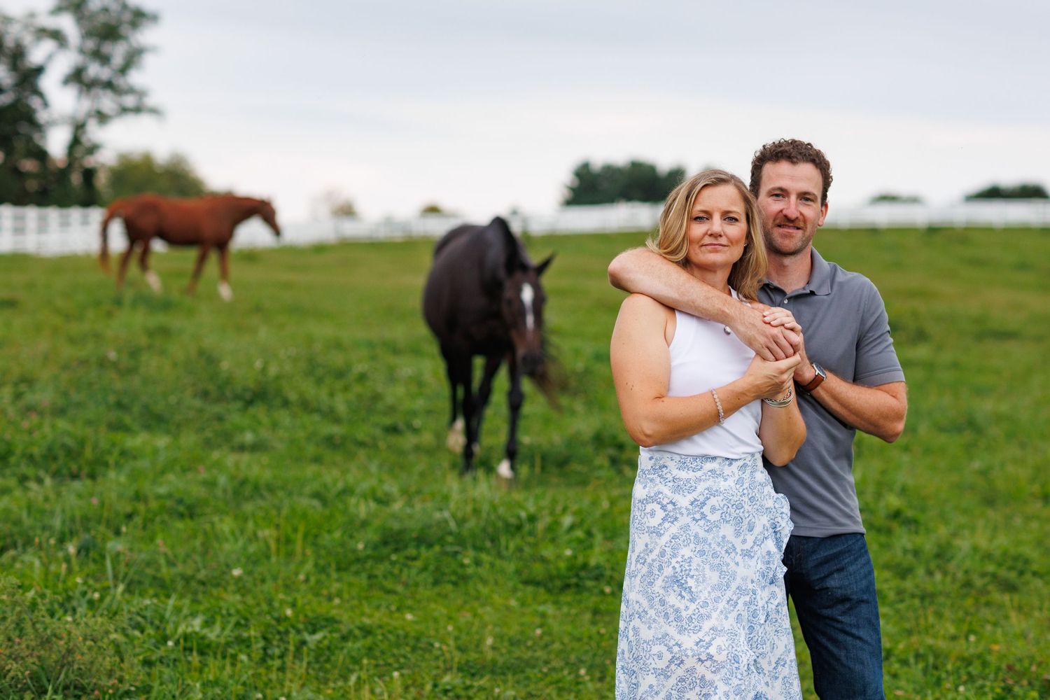 Happy couple embracing in a green pasture with two horses grazing behind them at golden hour.