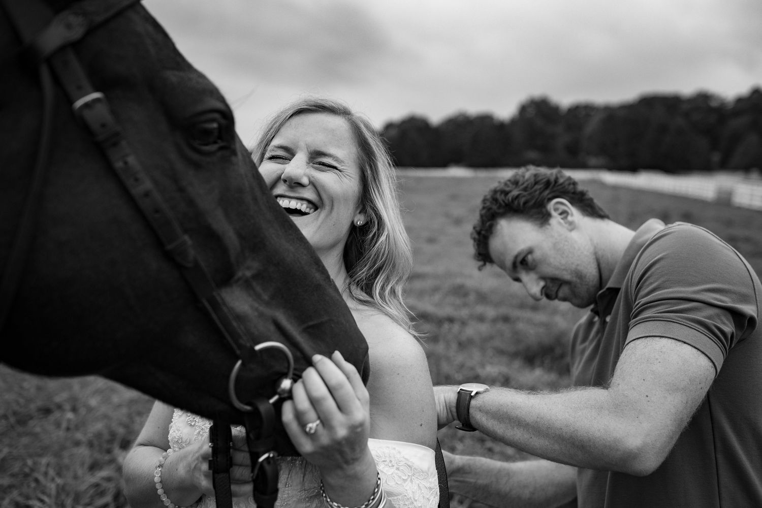 Black and white photo of a laughing couple with a dark horse in a field.