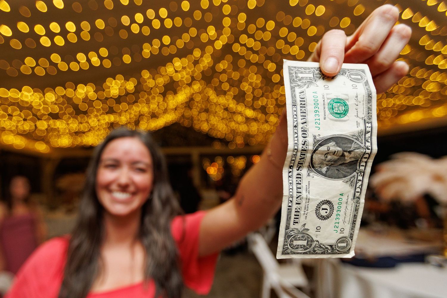Dollar bills being held up during a lively wedding dance celebration under golden string lights.