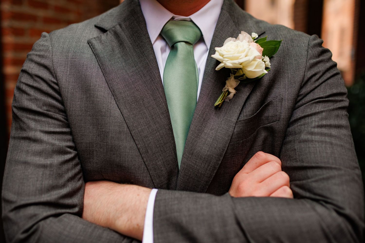 Groom in gray suit with mint green tie and white rose boutonniere, arms crossed at wedding.