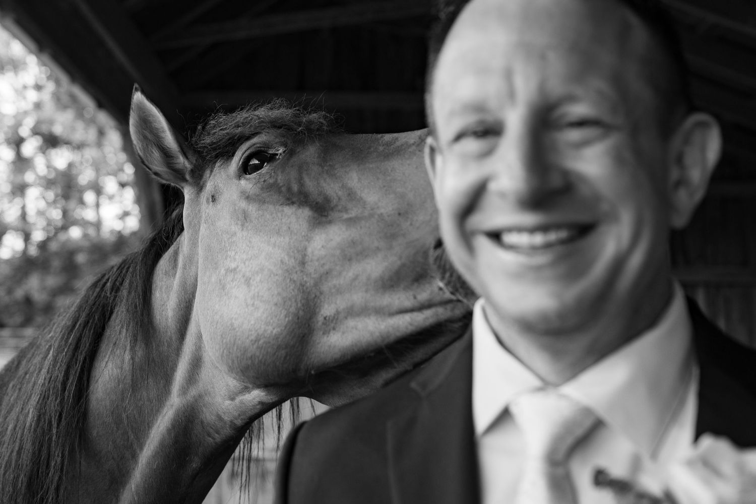 Black and white photo of a smiling groom in a suit being nuzzled by a horse outdoors.