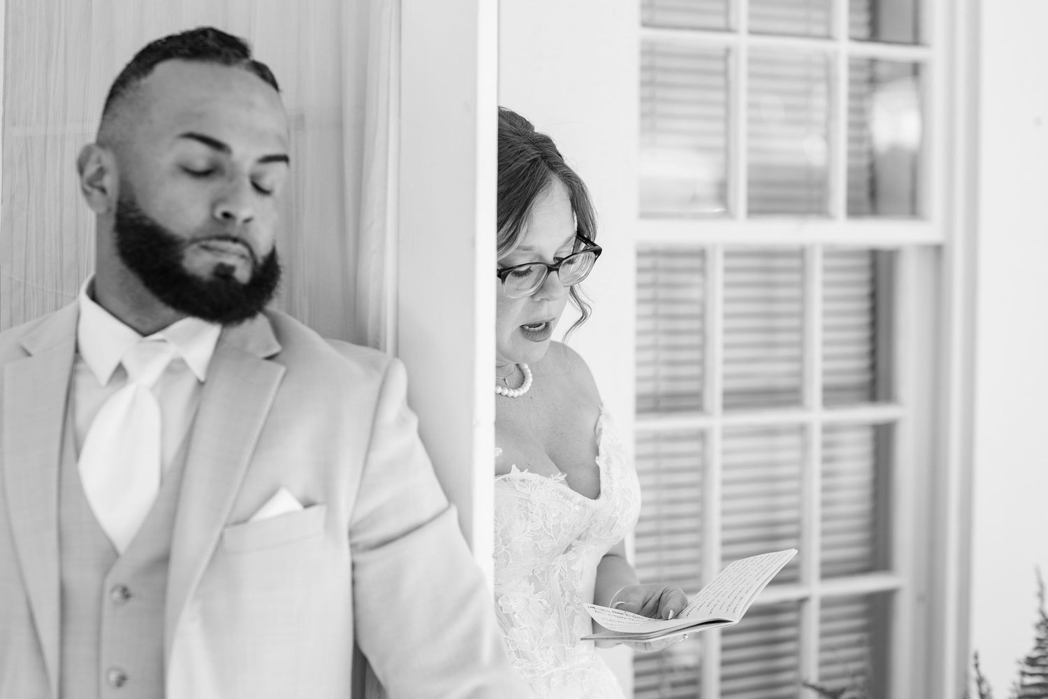 Black and white photo of groom standing against wall as bride reads vows behind a glass door.