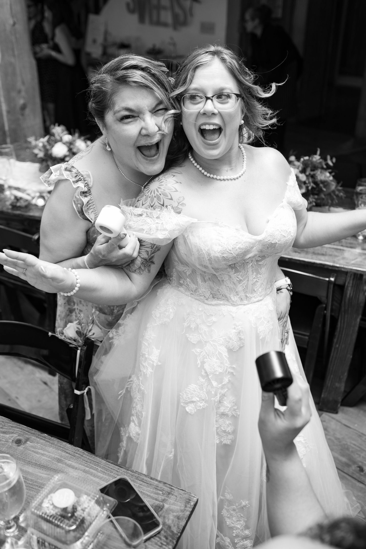 Two women in wedding attire laughing and embracing joyfully at wedding reception.