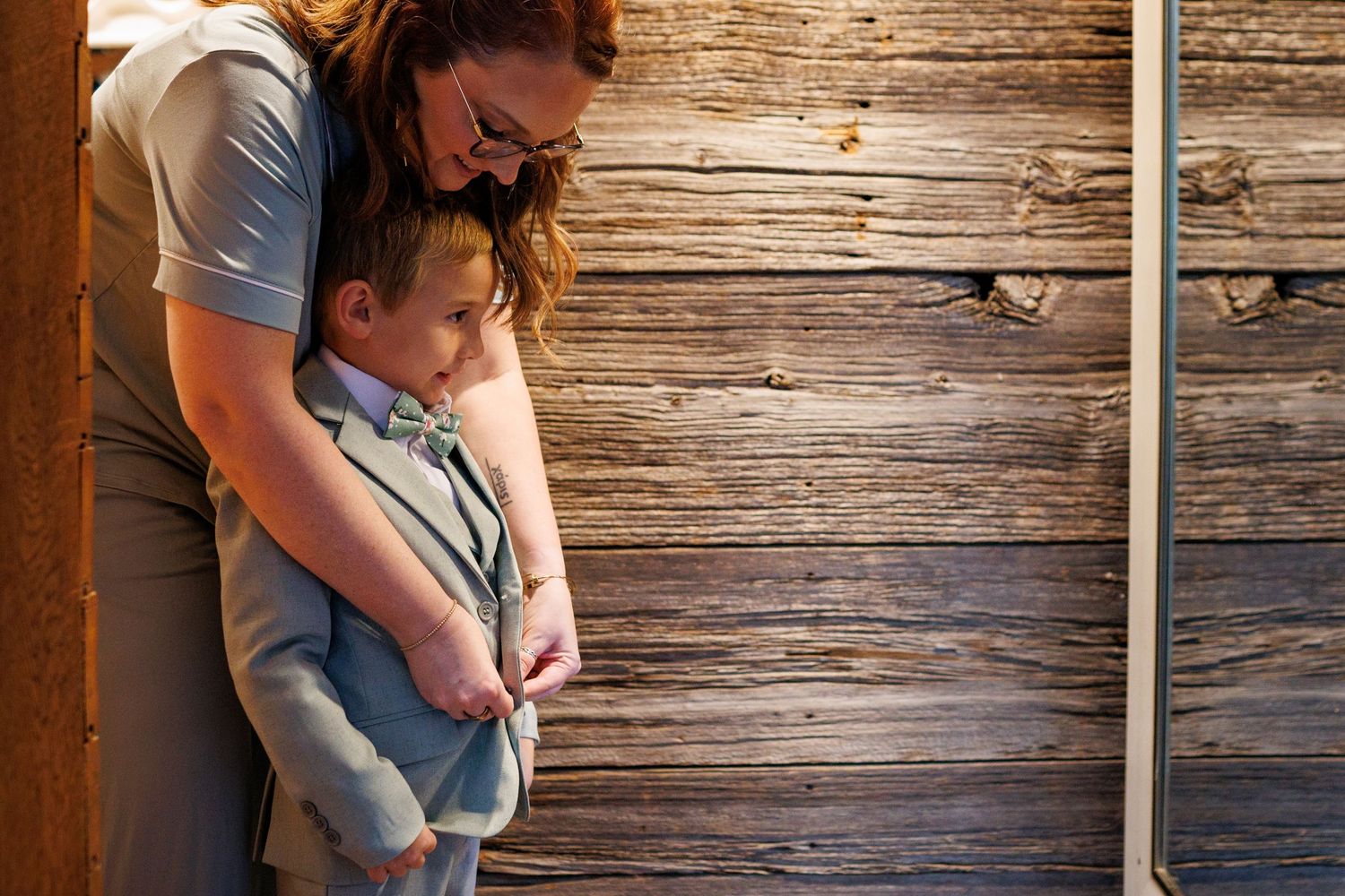 A mother tenderly embraces her young son in a bow tie against a rustic wooden wall backdrop.