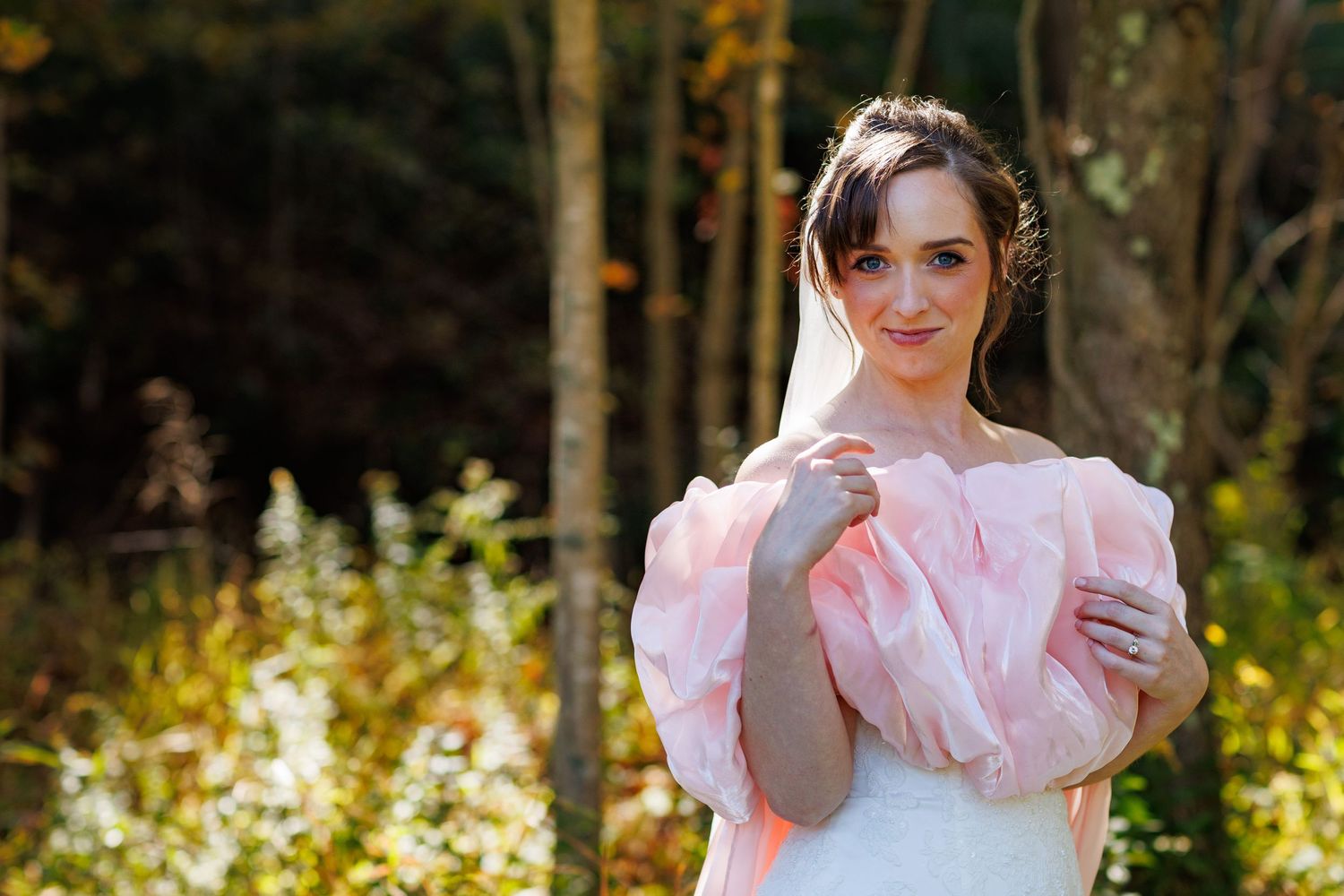 Bride in pink off-shoulder gown poses gracefully outdoors with autumn foliage background.