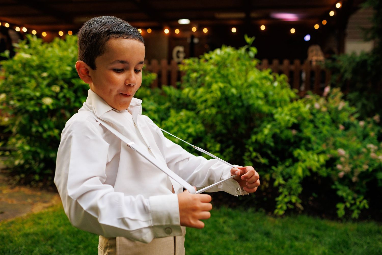 Young ring bearer in white shirt and tan suspenders adjusting his tie in a garden wedding setting.