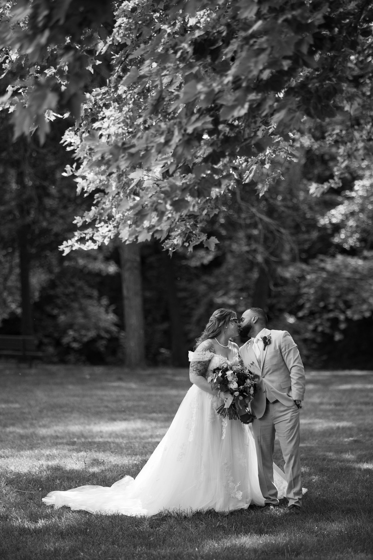 Black and white photo of bride and groom sharing a kiss under a large tree in a lush garden setting.
