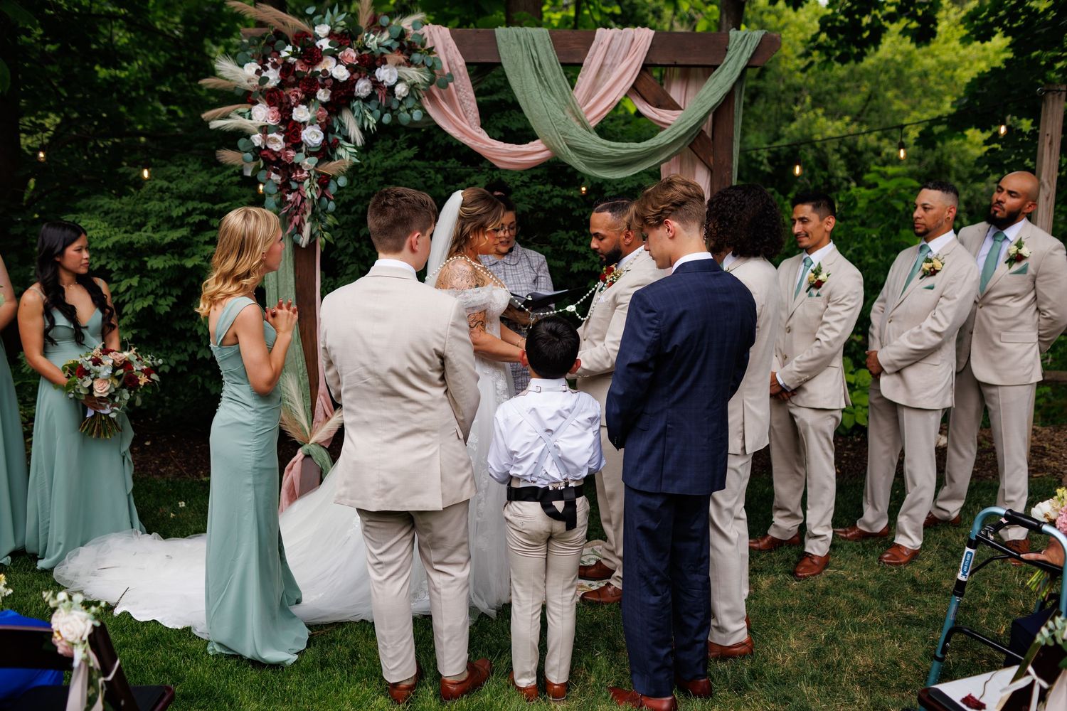 Outdoor wedding ceremony under floral arch with bride, groom, officiant, and wedding party in sage green and beige attire.