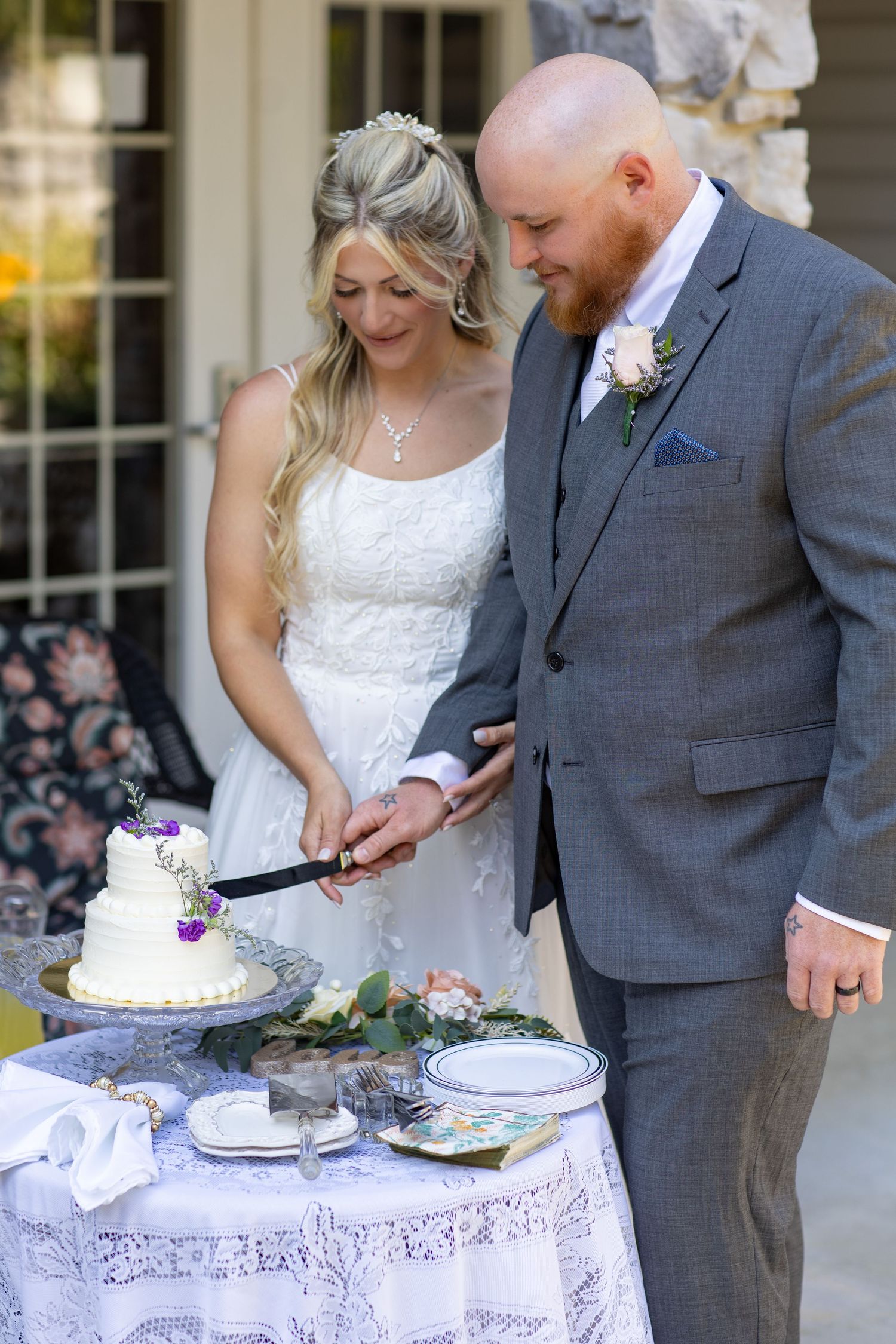 A couple in wedding attire cuts into their elegant white wedding cake decorated with purple flowers at their reception.
