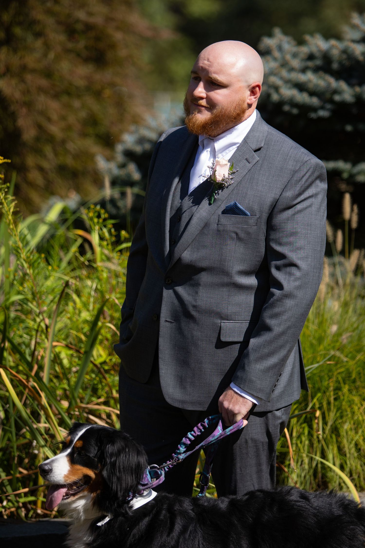 A person in a gray suit stands outdoors in tall grass with spruce trees visible in the background.