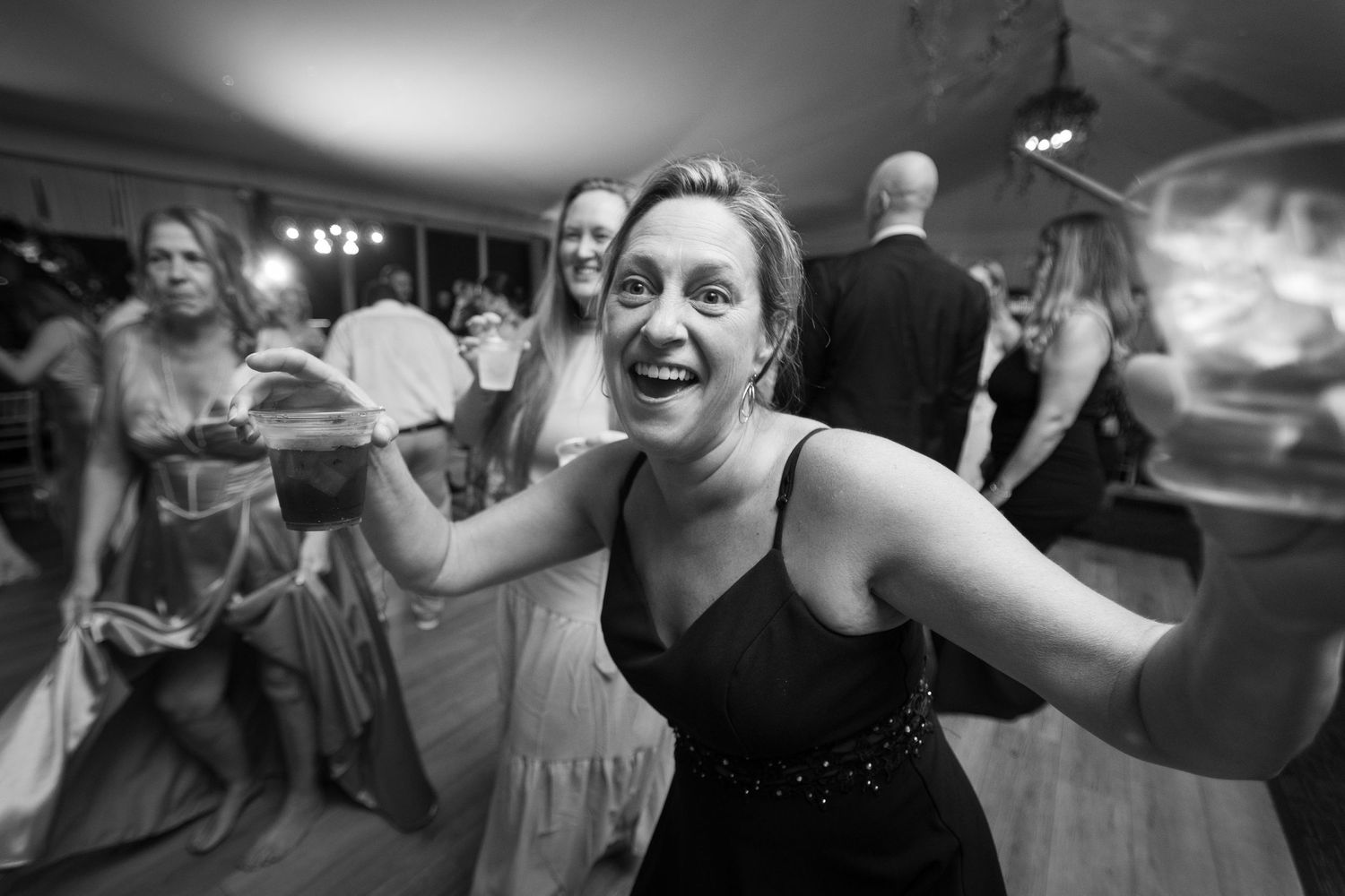 A joyful dancer in a black dress spreads their arms wide on a lively dance floor at a celebration.