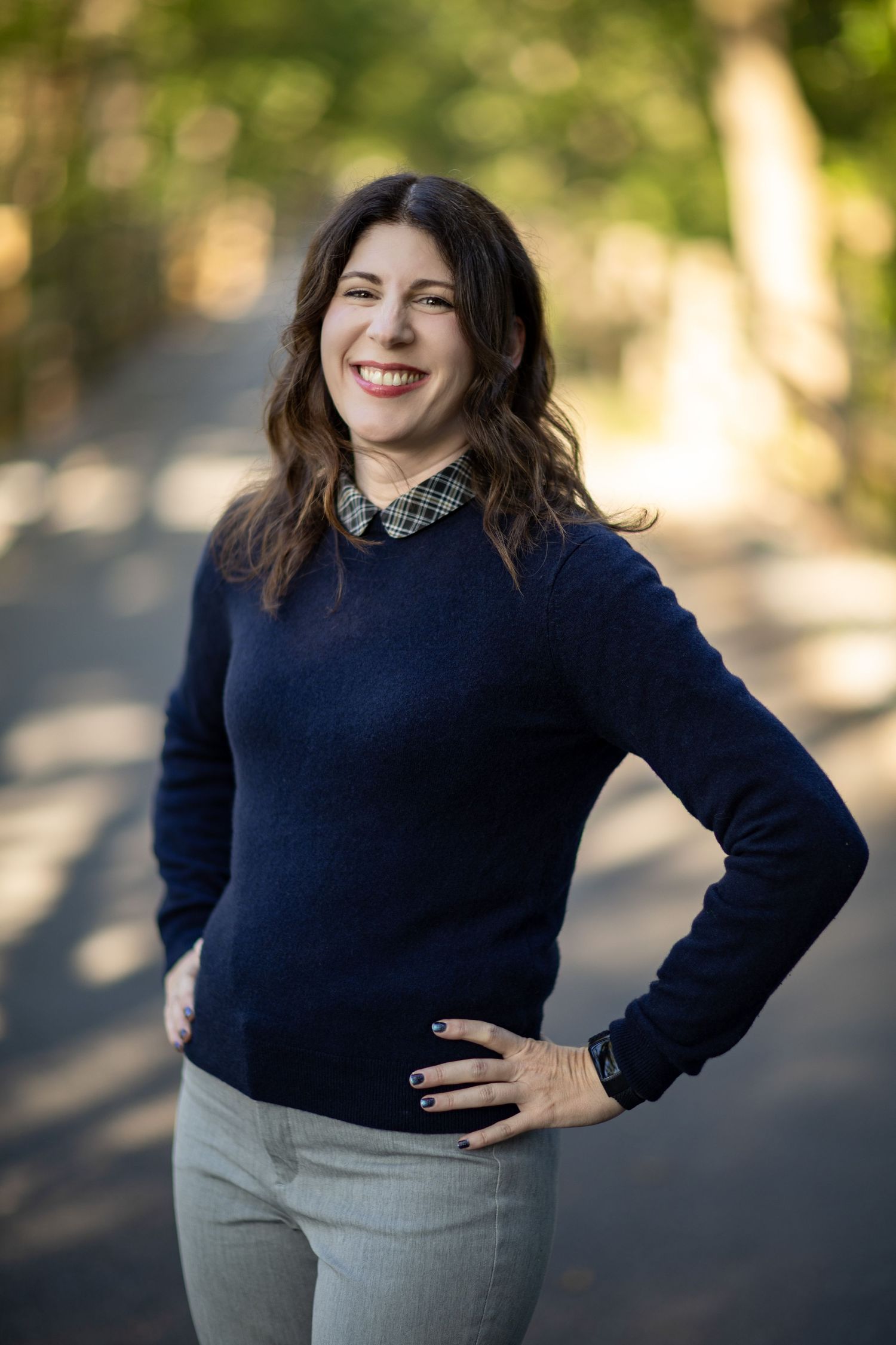 A woman in a navy sweater and gray skirt poses outdoors with natural lighting and bokeh background.