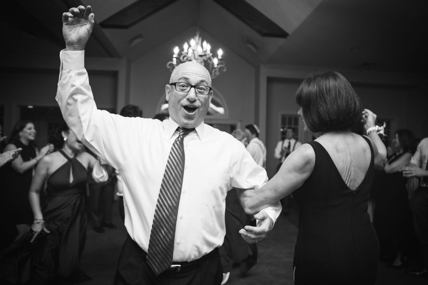 Person wearing glasses and tie raises arm while dancing at a formal celebration.