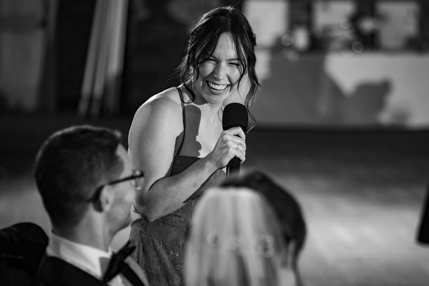 Black and white photo of a woman smiling and speaking into a microphone at a wedding reception.